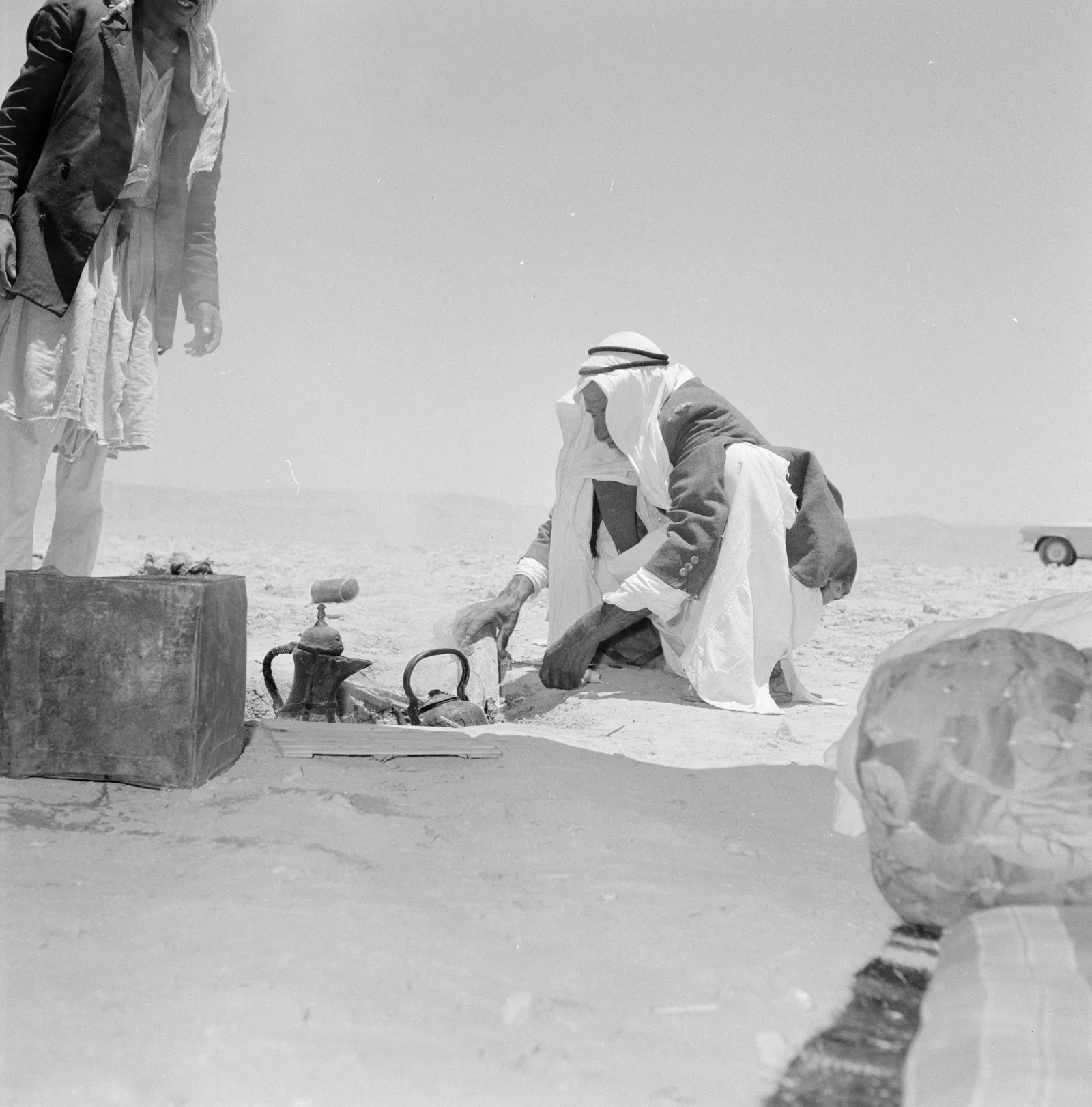 Photographie de deux hommes accroupis dans le désert autour d'un foyer de sable chaud, où sont enfoncées des cafetières à long bec et des ustensiles servant à préparer le café.