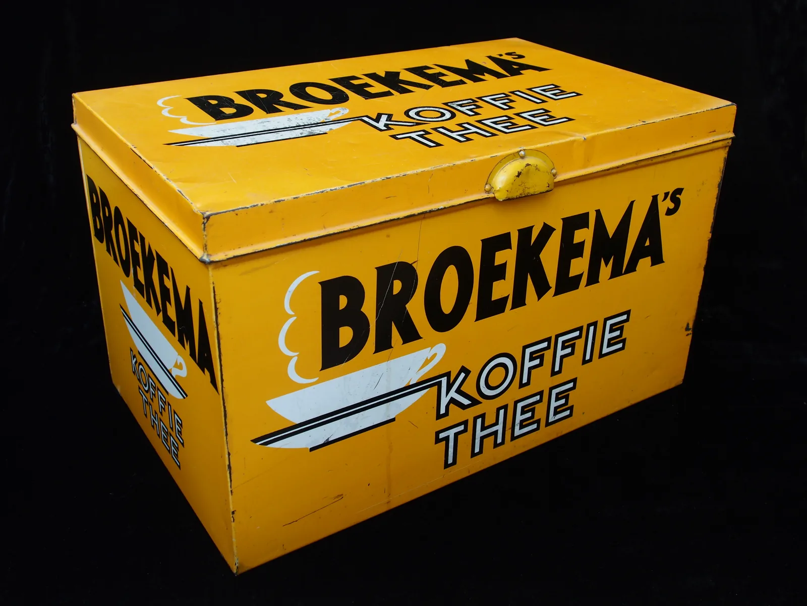 Photographie d'une grande boîte de comptoir en tôle jaune à couvercle et fermoir, dont les faces portent le nom de la maison Broekema en lettres bleu foncé et les mots café et thé.
