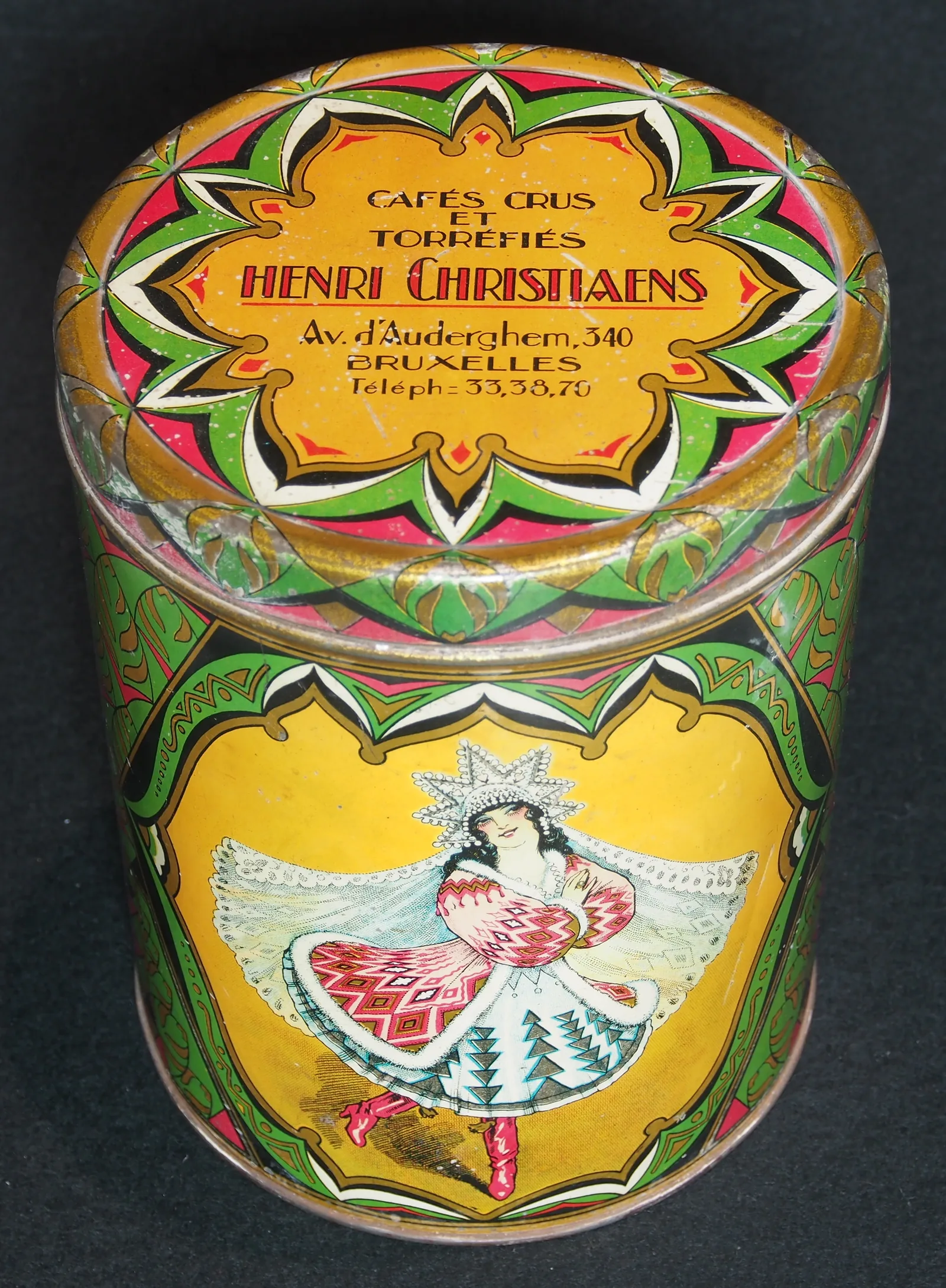 Photographie d'une boîte cylindrique de tôle peinte : le couvercle porte en couronne le nom de la maison Henri Christiaens, la mention cafés crus et torréfiés et l'adresse bruxelloise, la paroi étant décorée d'un oiseau stylisé sur fond jaune.