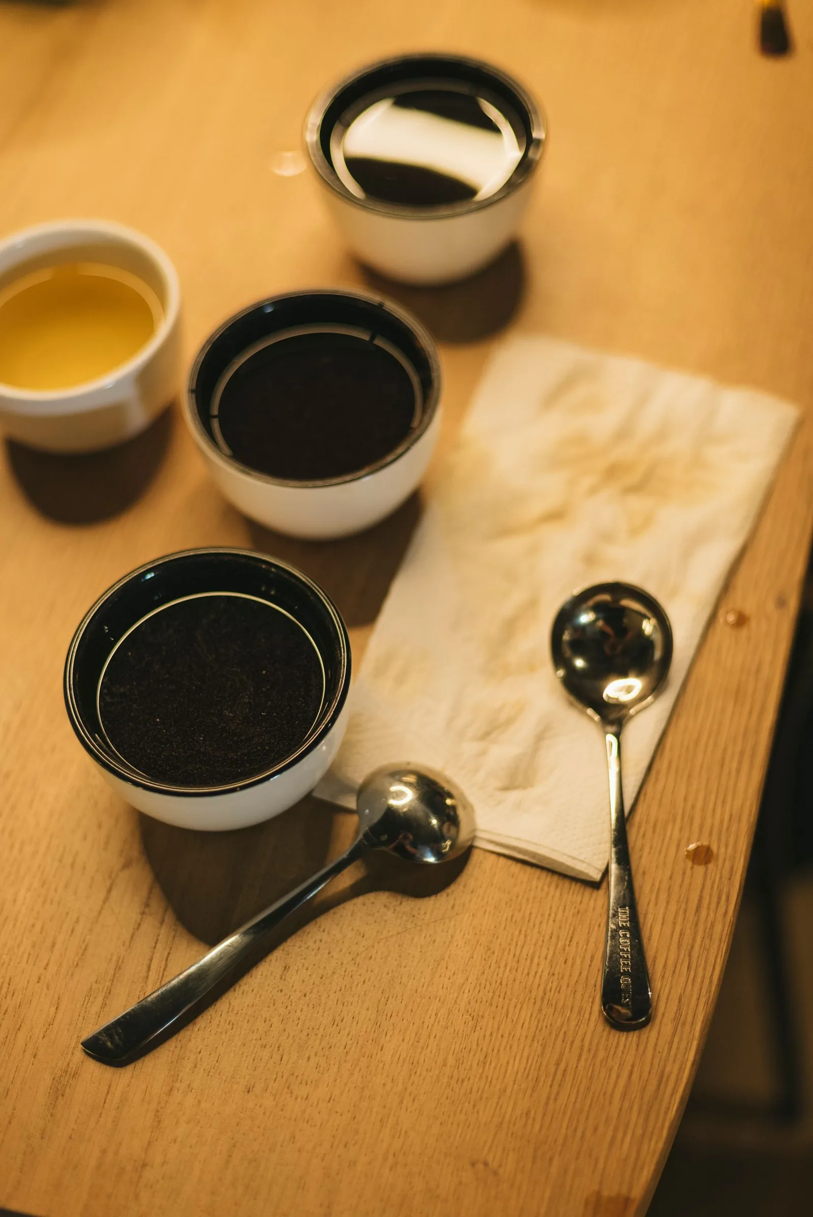 Sur une table de bois, trois bols contiennent la mouture sèche et un quatrième un liquide clair ; deux cuillères de dégustation sont posées à côté.