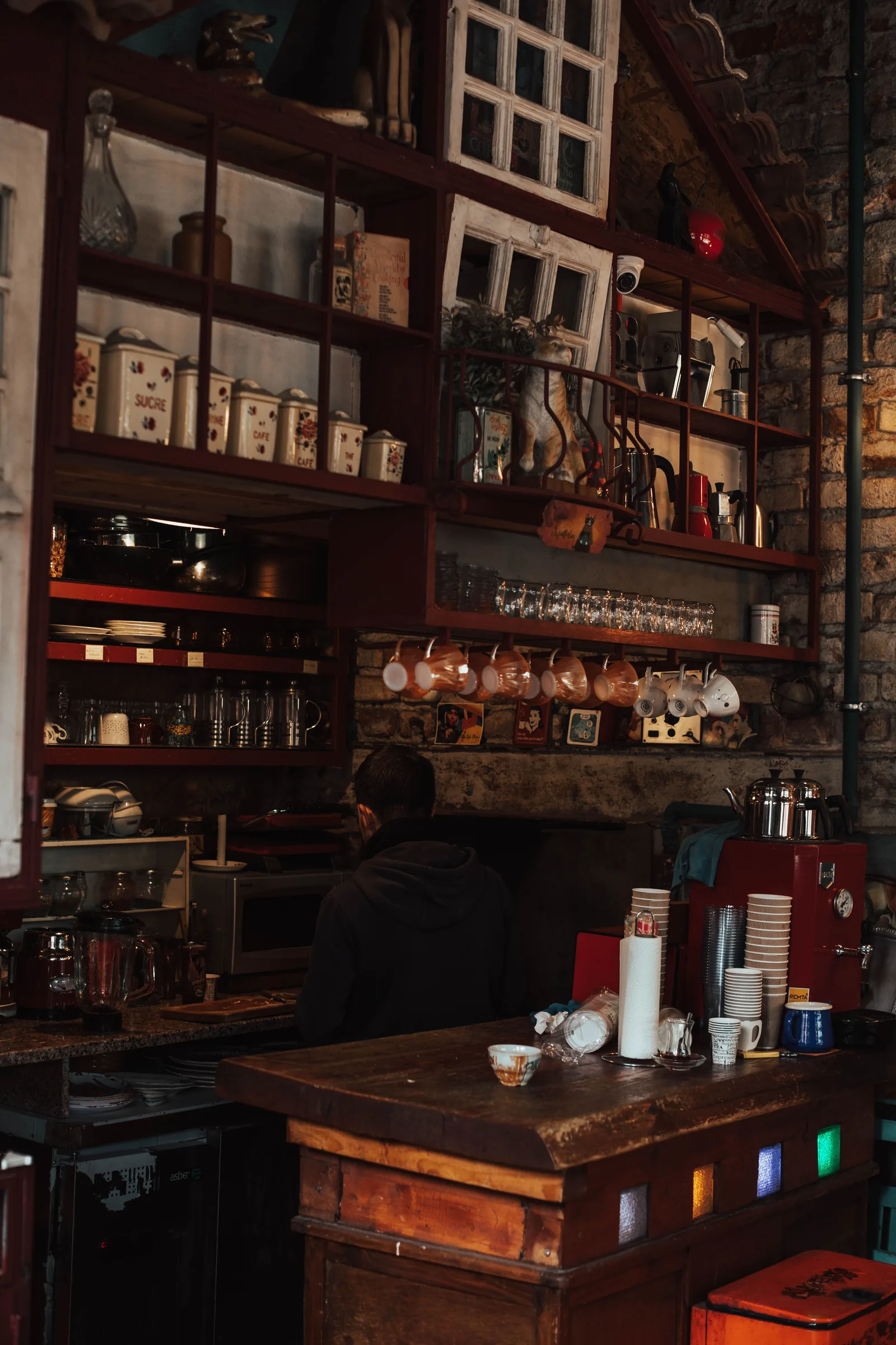 Un homme vu de dos derrière le comptoir encombré d'un café de quartier, entouré de boîtes à sucre et à café, de tasses dépareillées suspendues et de gobelets empilés