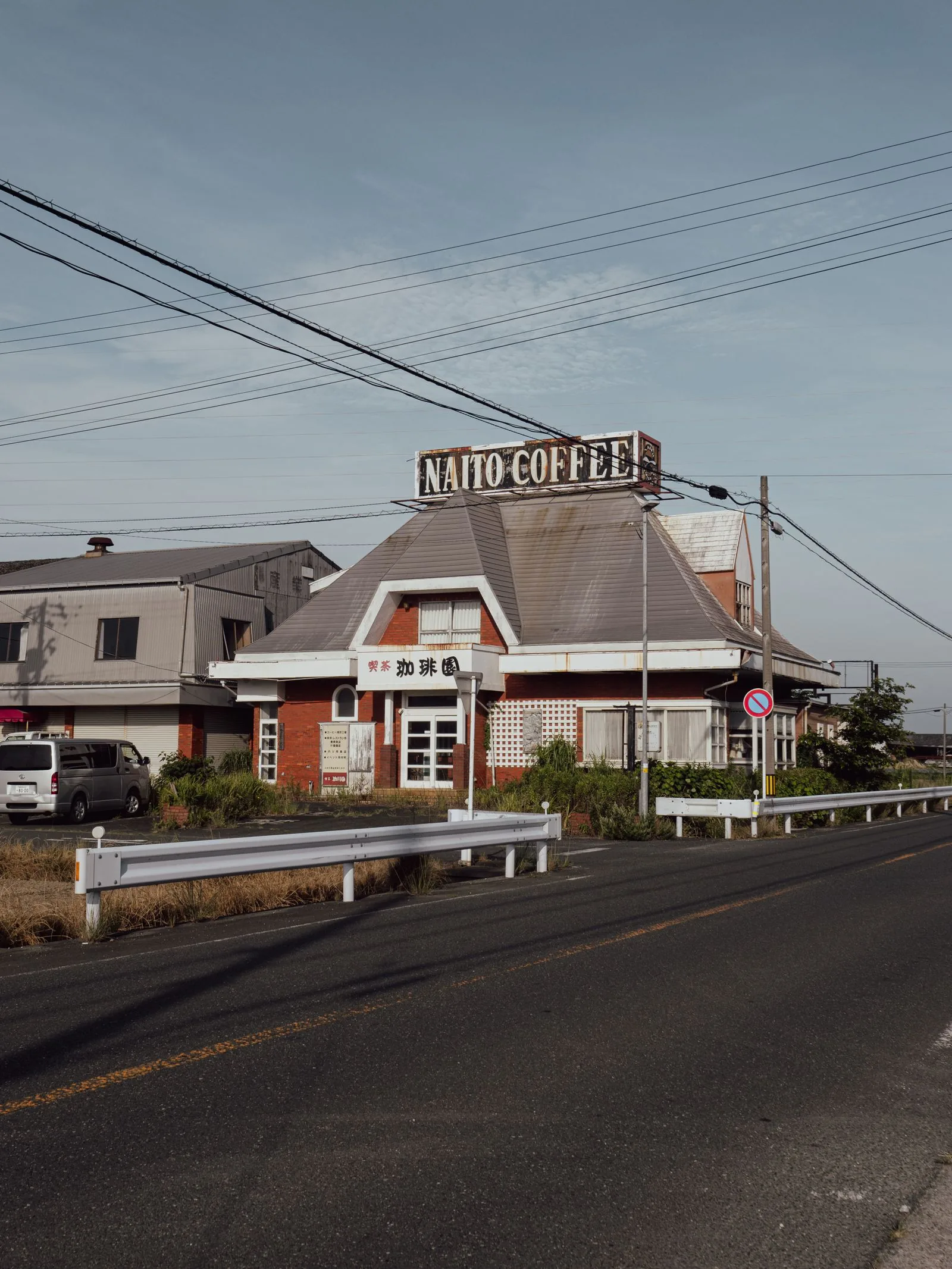 Bâtiment bas au toit pentu au bord d'une route à deux voies, enseigne « NAITO COFFEE » sur le faîtage, fils électriques au-dessus de la chaussée.