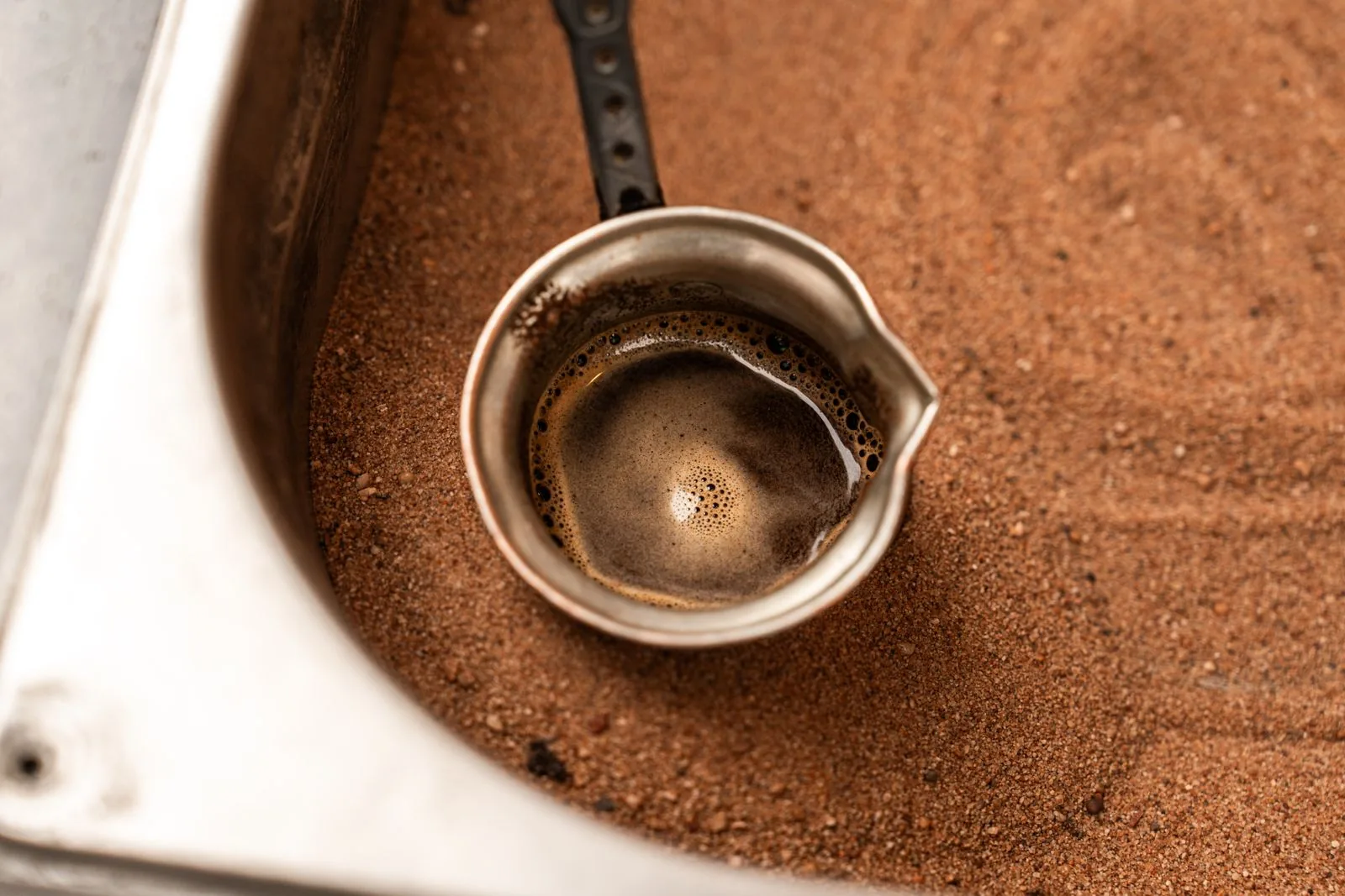 Vue plongeante sur un pot de métal à bec verseur planté dans du sable brun, rempli d'un café mousseux.