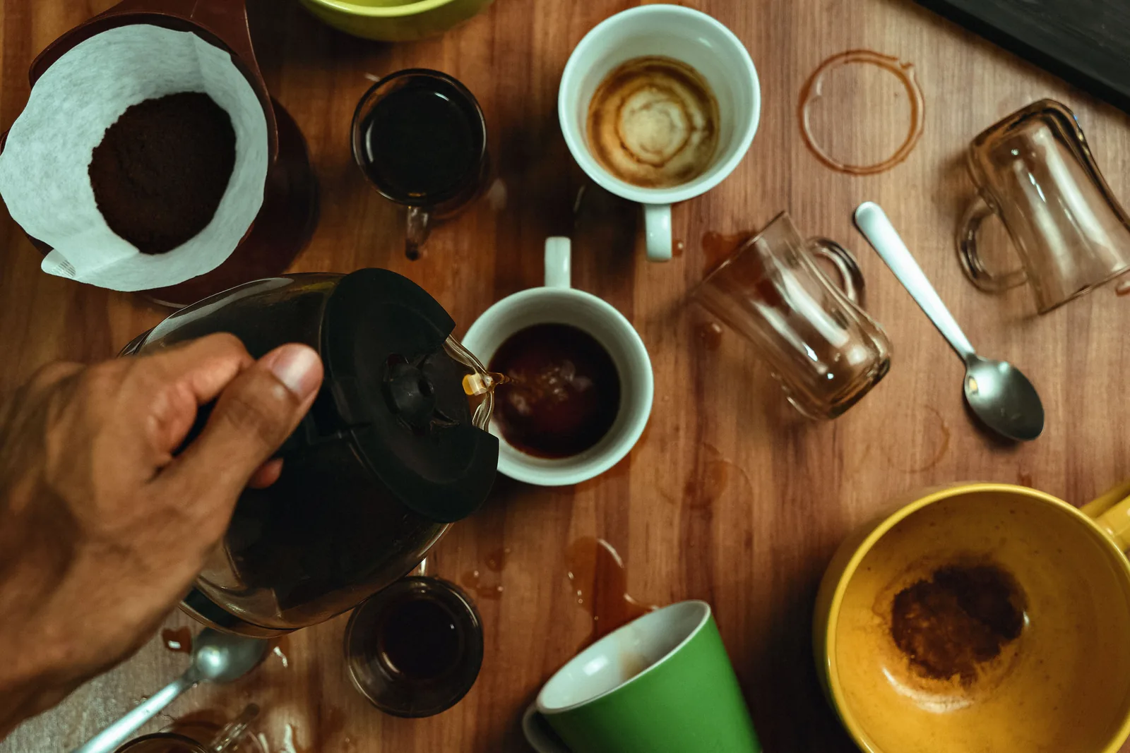 Une main verse du café depuis une cafetière dans une tasse blanche entourée de plusieurs autres tasses et verres dépareillés sur une table en bois tachée de café
