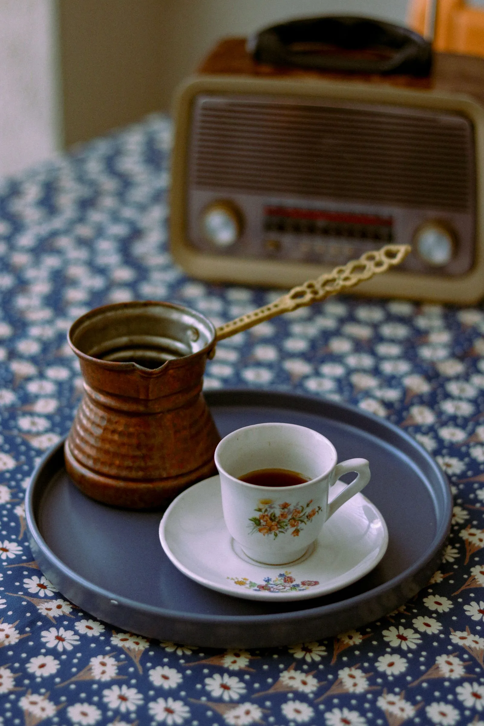 Petit pot de cuivre à long manche et tasse à fleurs sur sa soucoupe, posés sur un plateau gris, nappe fleurie et poste de radio ancien derrière.