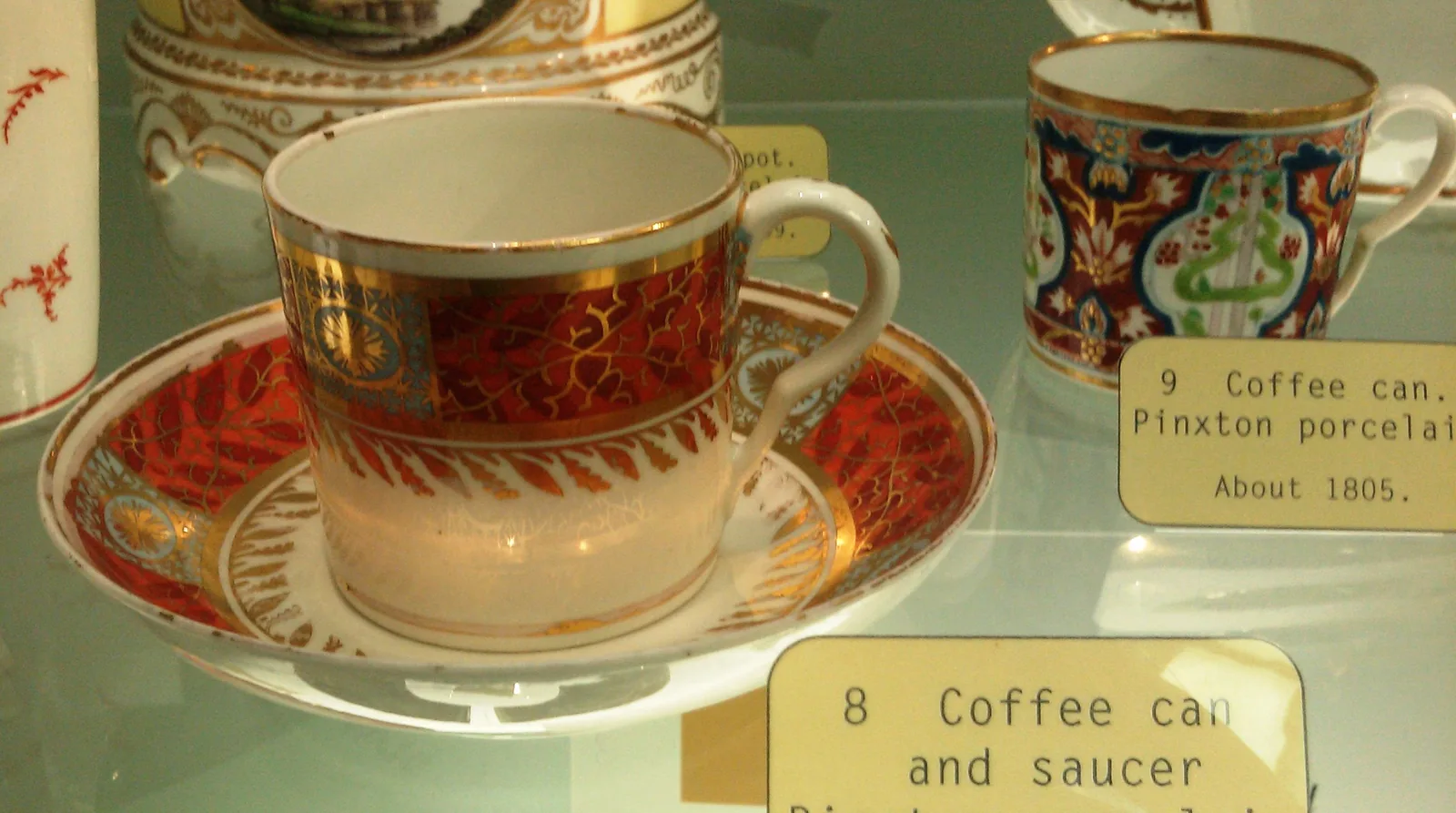 Photographie prise dans une vitrine de musée montrant des gobelets à café de porcelaine de Pinxton avec leurs soucoupes, décorés de bandeaux rouges, verts et dorés, accompagnés de leurs cartels imprimés.