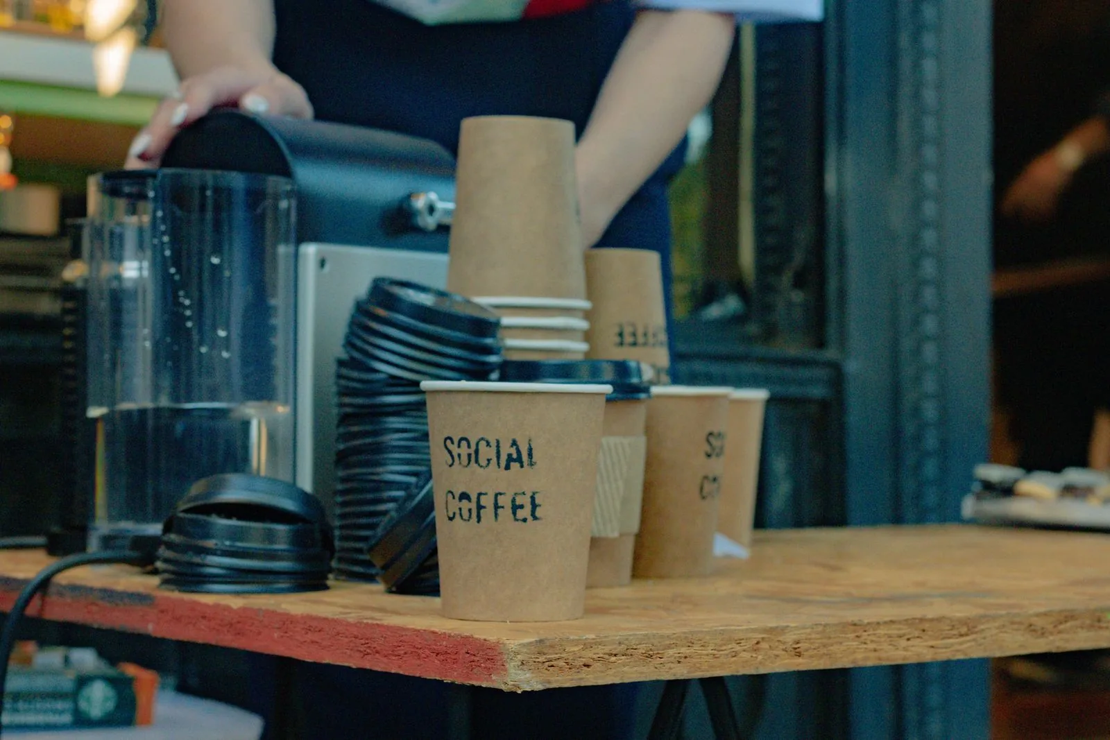 Gobelets de carton marqués « SOCIAL COFFEE » et piles de couvercles bleus sur un plateau de bois, mains d'un serveur au second plan.