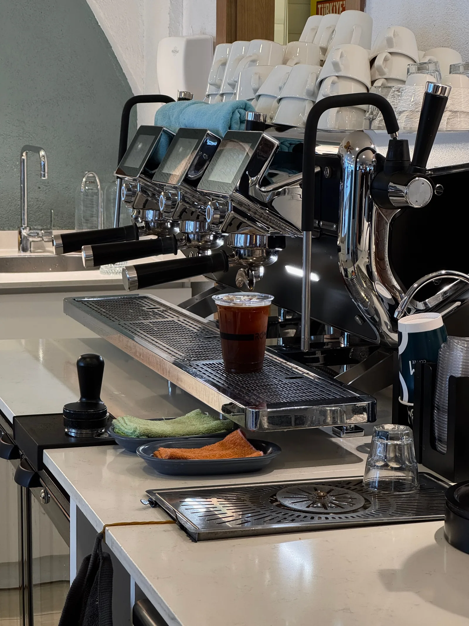Gros plan sur un comptoir de café avec une machine à espresso professionnelle à trois groupes, des tasses blanches empilées, un tasseur noir posé au premier plan et un café glacé sous un porte-filtre