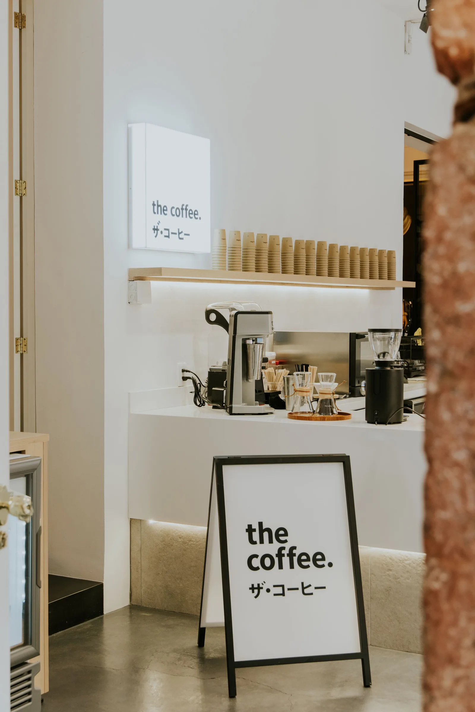 Un comptoir de café blanc et épuré, vu depuis la salle, avec une machine, un moulin, des filtres Chemex et une pile de gobelets en carton sous une enseigne lumineuse