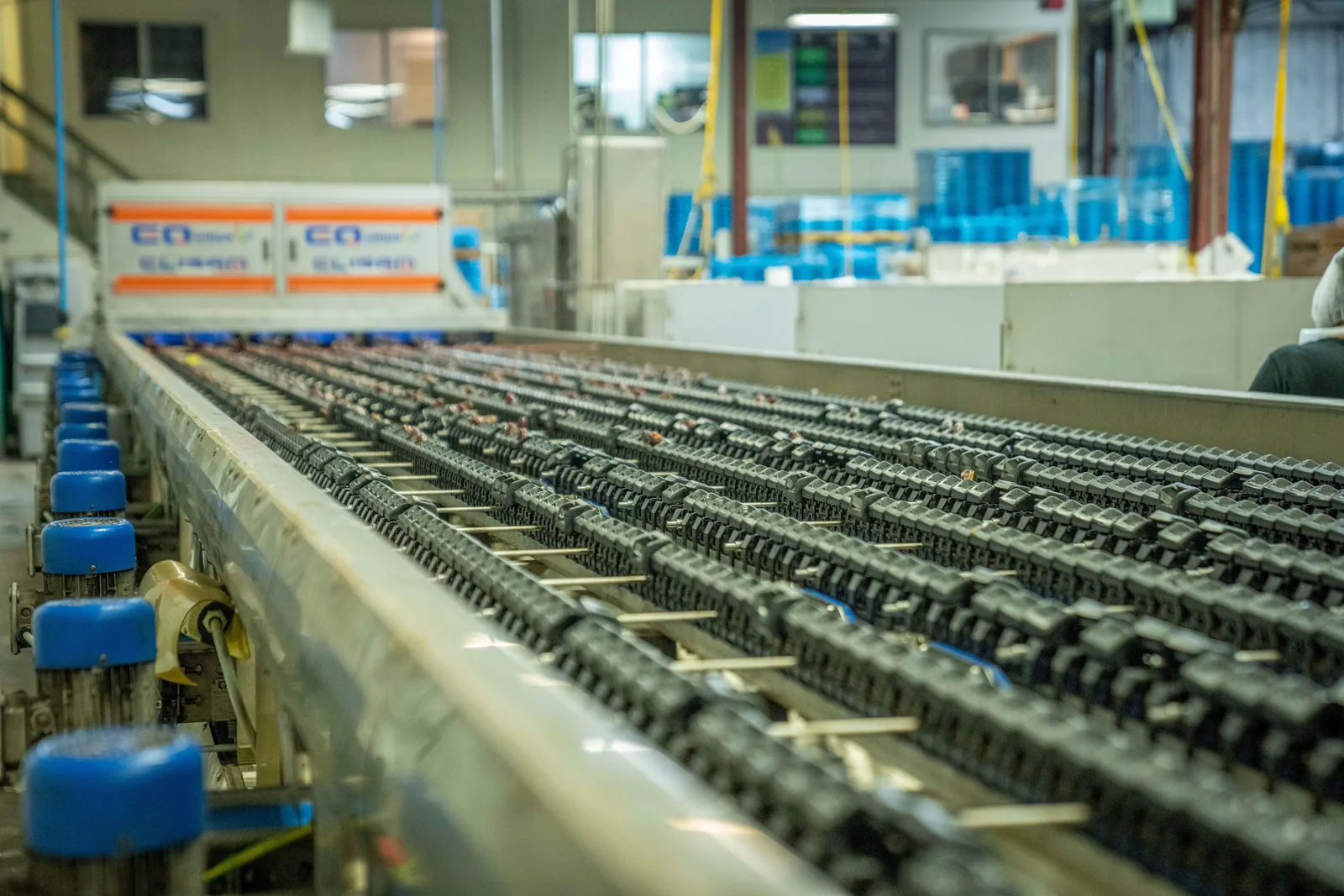 Une longue ligne de convoyeur mécanique traversant un atelier industriel, avec des moteurs bleus au premier plan et une silhouette floue au fond, sans visage reconnaissable