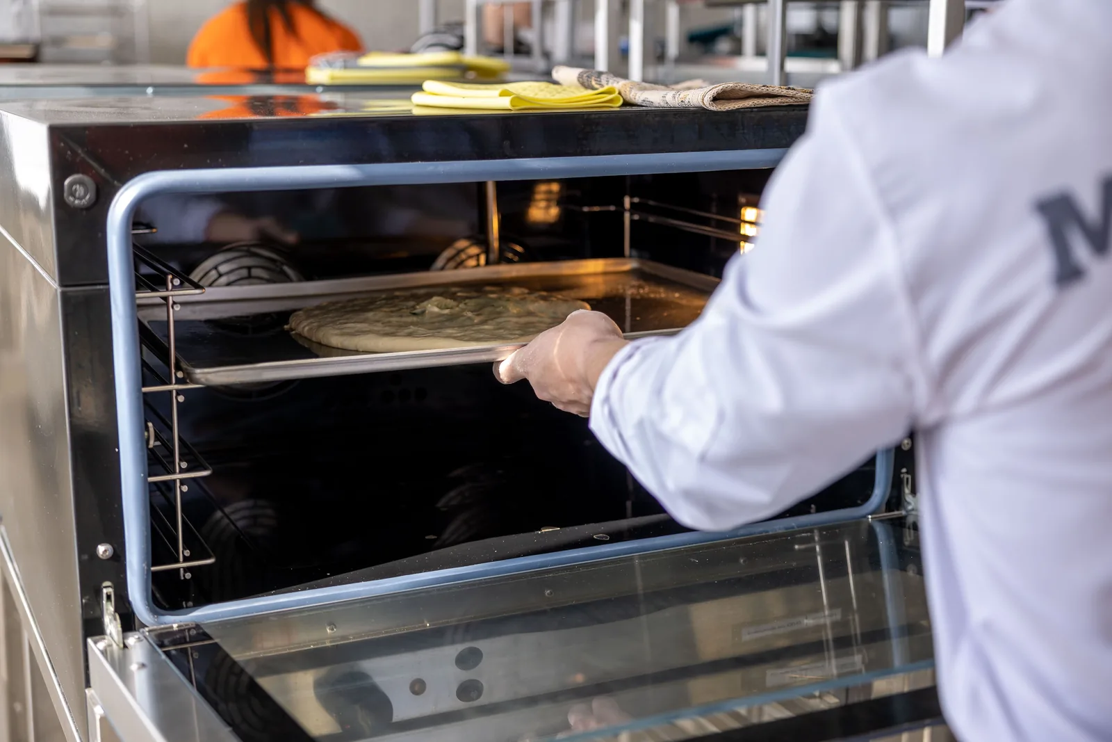 Un cuisinier vu de dos glisse une pâte à pizza crue sur une plaque à l'intérieur d'un four professionnel ouvert, ventilateur de convection visible à l'intérieur