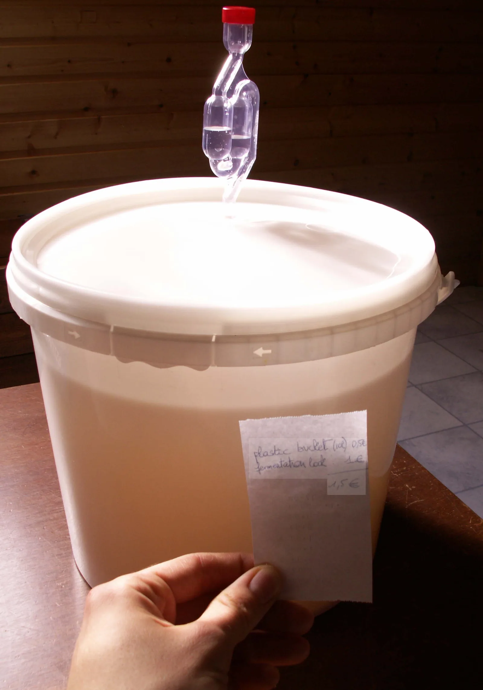 Un bidon de fermentation en plastique alimentaire fermé par un couvercle percé d'un barboteur à eau rempli de liquide, une main tenant une étiquette de prix devant