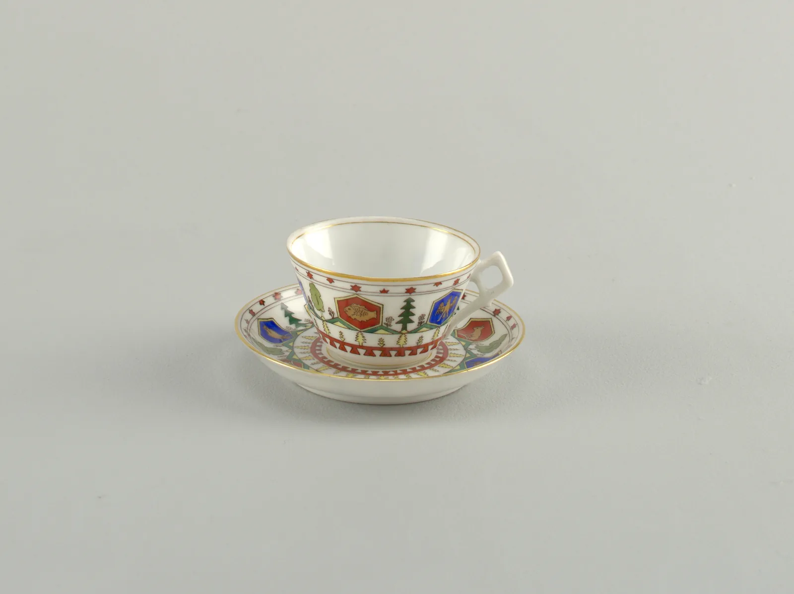 Photographie d'une petite tasse à café russe à paroi évasée et de sa soucoupe, décorées de cartouches hexagonaux rouges et bleus à figures animales, d'arbres verts et d'etoiles.