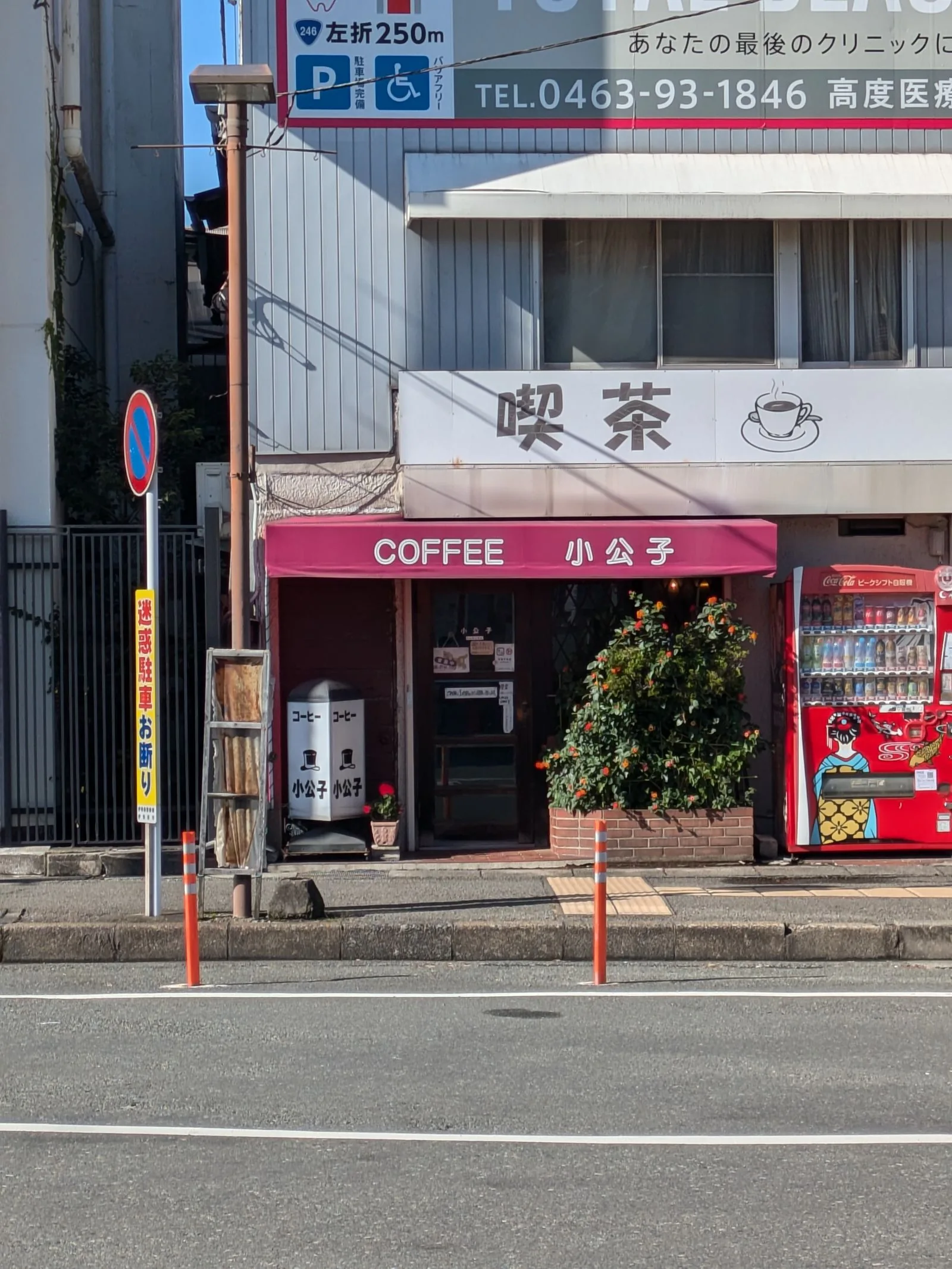 Petite façade de rue avec une enseigne blanche portant les caractères « 喫茶 » et une tasse dessinée, au-dessous une enseigne rose « COFFEE », distributeurs automatiques à droite.