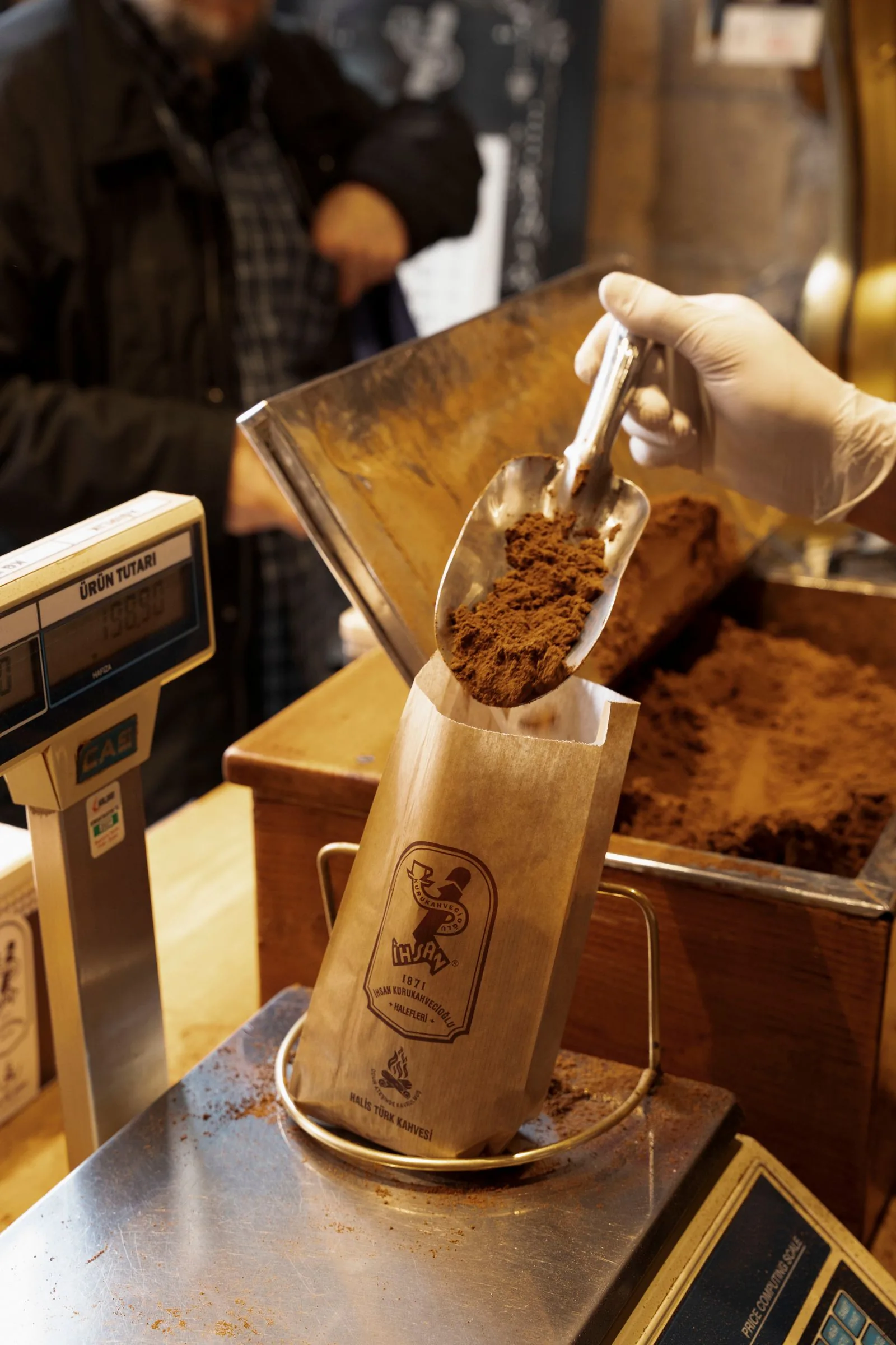 Main gantée versant du café moulu à la pelle dans un sachet de papier posé sur le plateau d'une balance de boutique.