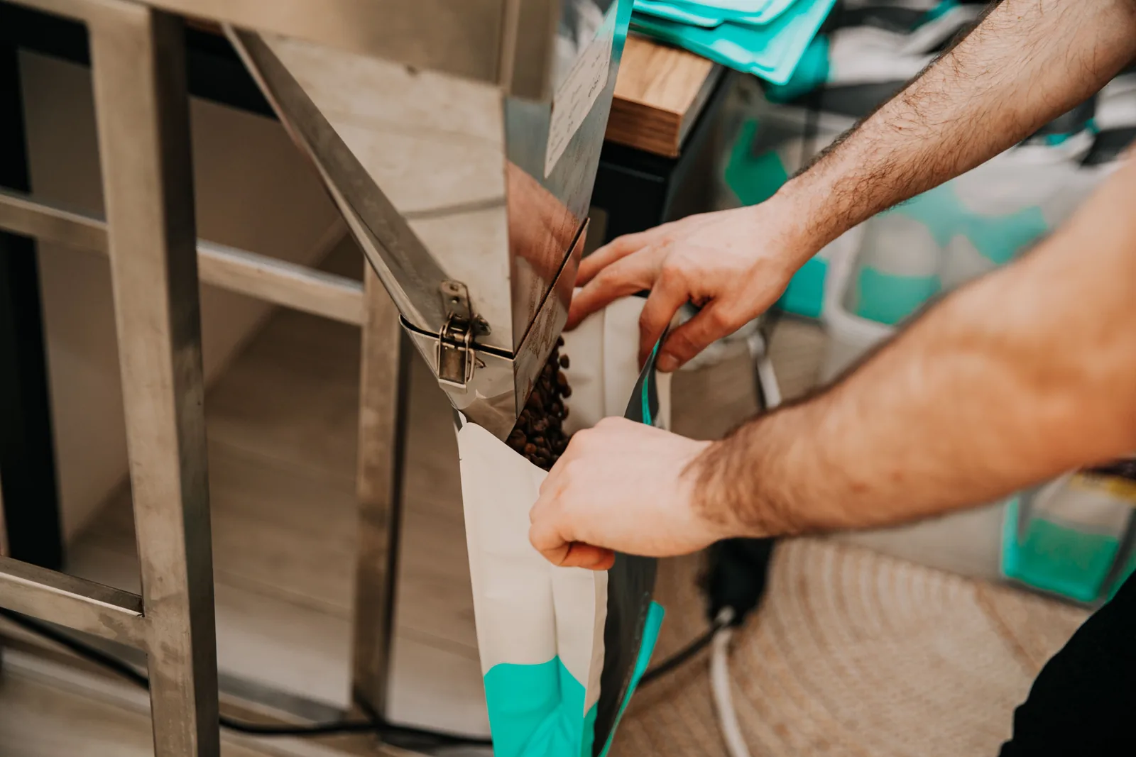 Des mains tiennent un sac ouvert sous une trémie métallique d'où s'écoulent des grains de café torréfiés