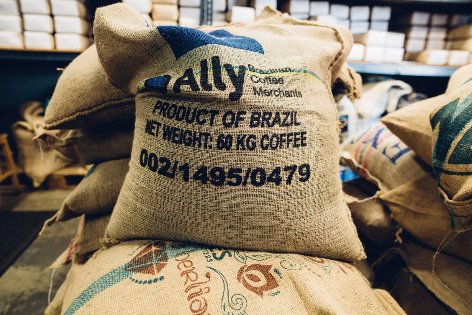 Sac de jute marqué « Product of Brazil, net weight 60 kg coffee » et d'un numéro de lot, posé sur d'autres sacs d'origines différentes.