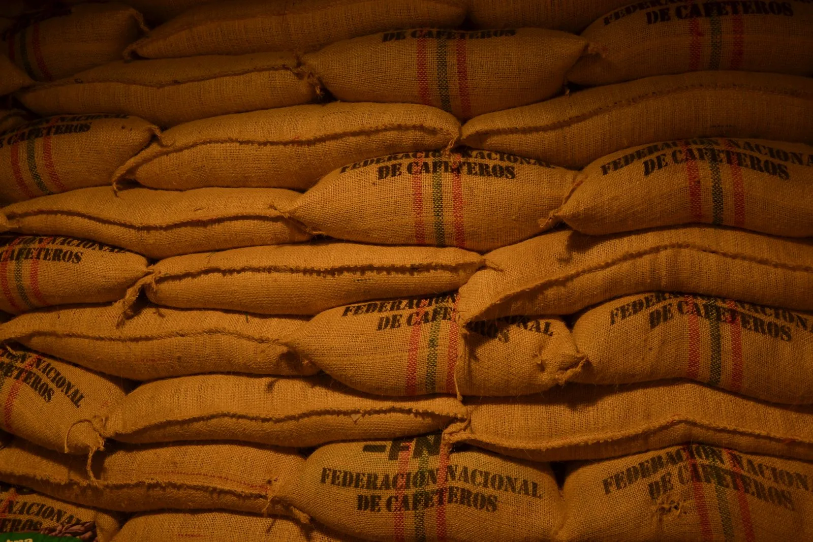Mur de sacs de jute empilés en quinconce, chacun portant la mention « Federación Nacional de Cafeteros » et deux liserés rouge et vert.