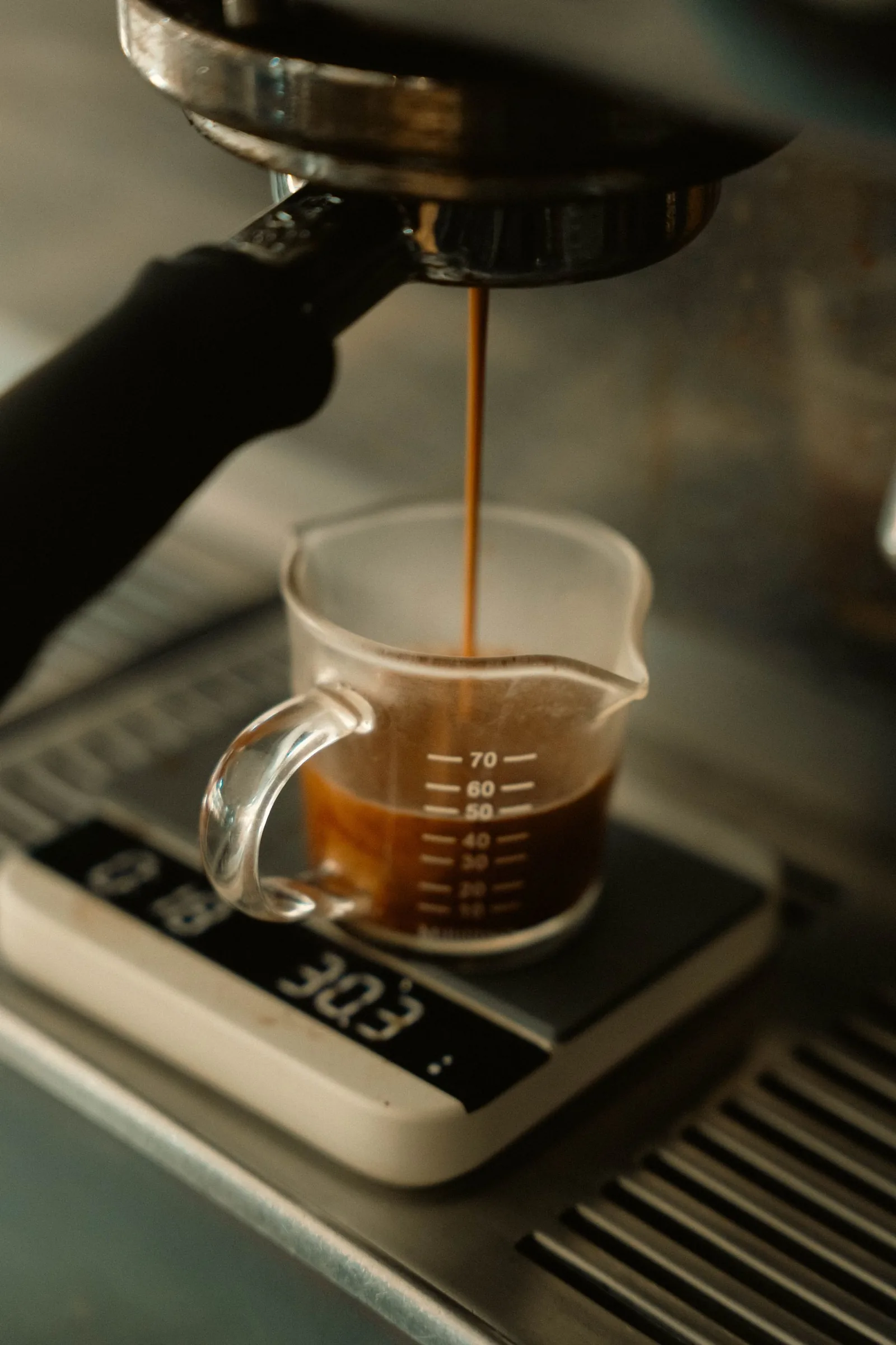 Le café coule du porte-filtre dans un petit verre gradué posé sur une balance électronique, contre le flanc chromé de la machine.