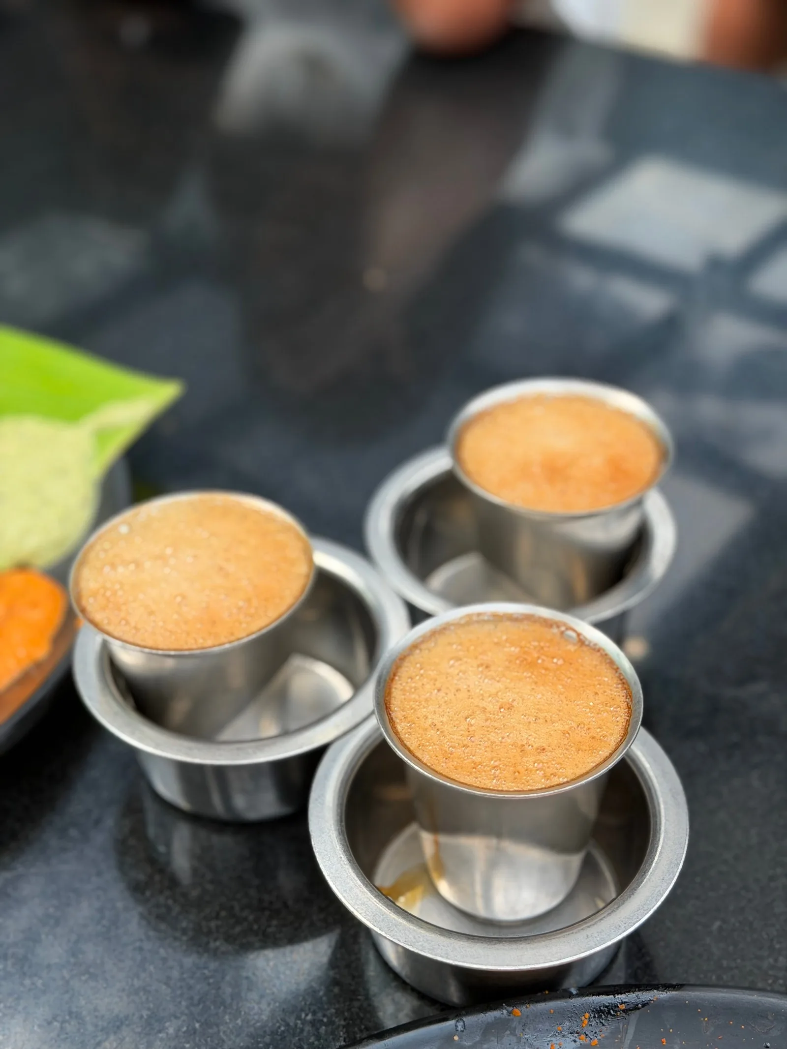 Trois gobelets d'acier dans leurs coupelles, coiffés d'une mousse épaisse, posés sur un plan de travail gris.