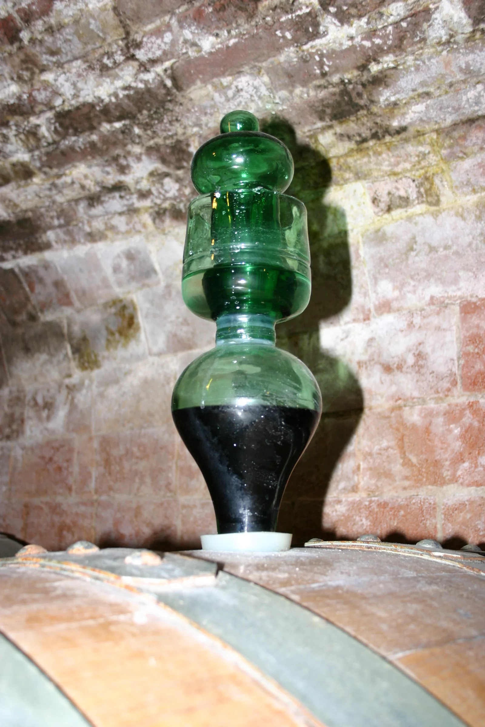 Un barboteur en verre vert vissé sur le bondon d'un fût de bois, dans une cave voûtée en briques ; l'image montre une fermentation de vin, mais l'objet photographié, un barboteur sur cuve fermée, est identique à celui utilisé pour la fermentation du café