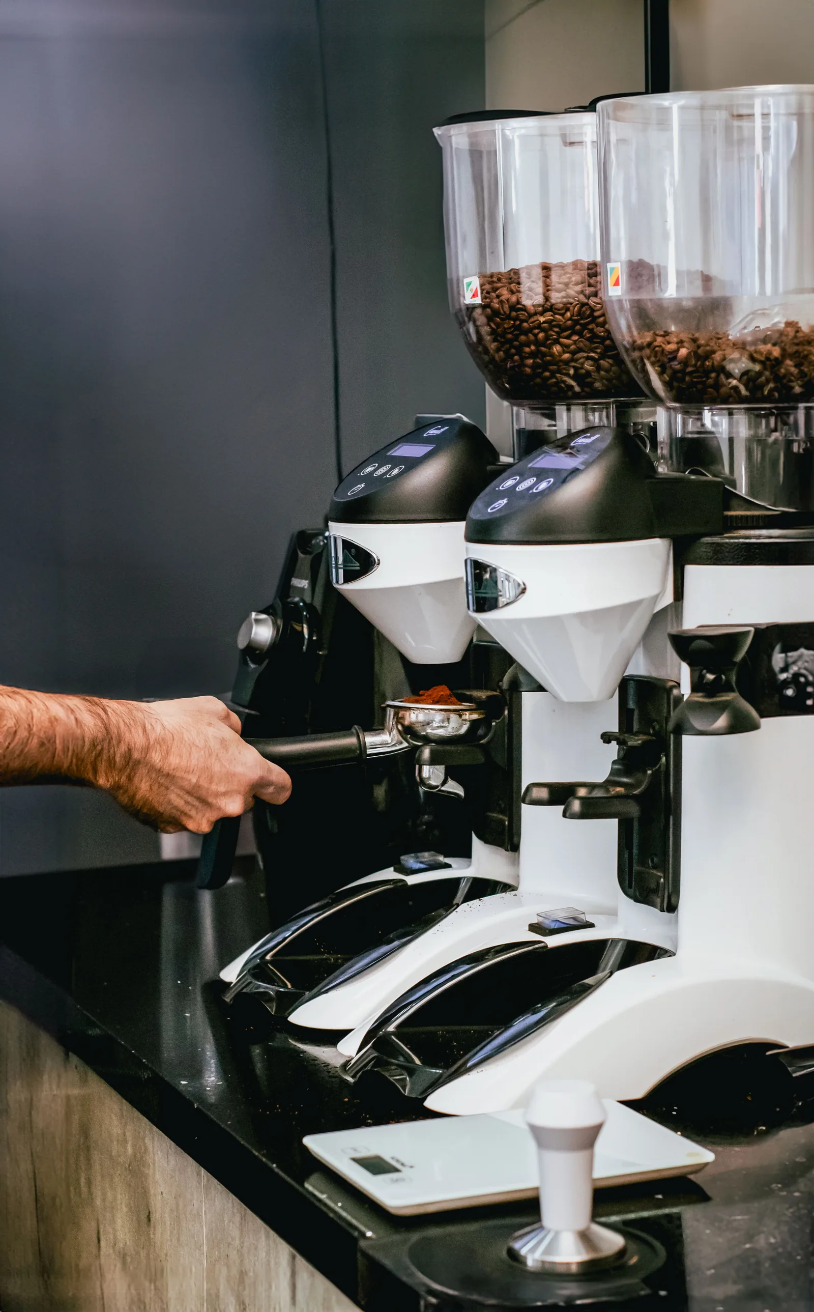 Une main tient un porte-filtre sous un moulin à café professionnel à deux trémies remplies de grains, avec un tasseur et une balance posés sur le comptoir noir