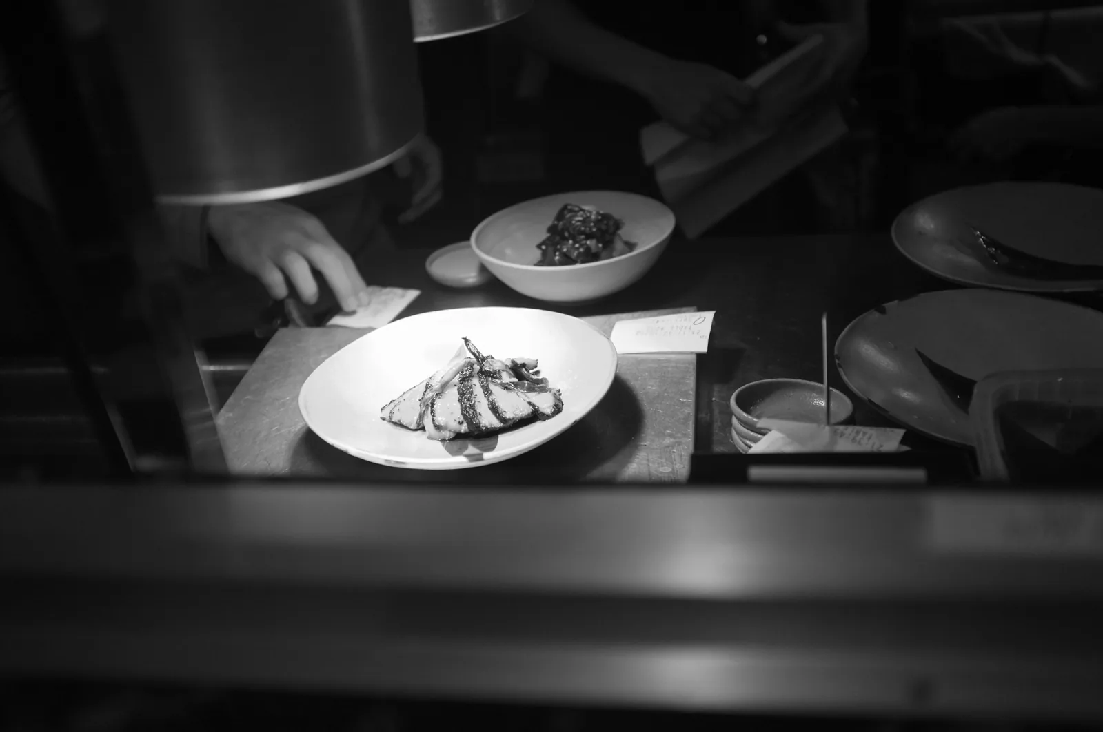 Photo en noir et blanc de deux assiettes de viande tranchée posées sur la passe d'une cuisine, avec des tickets de commande et des mains en arrière-plan, aucun visage visible