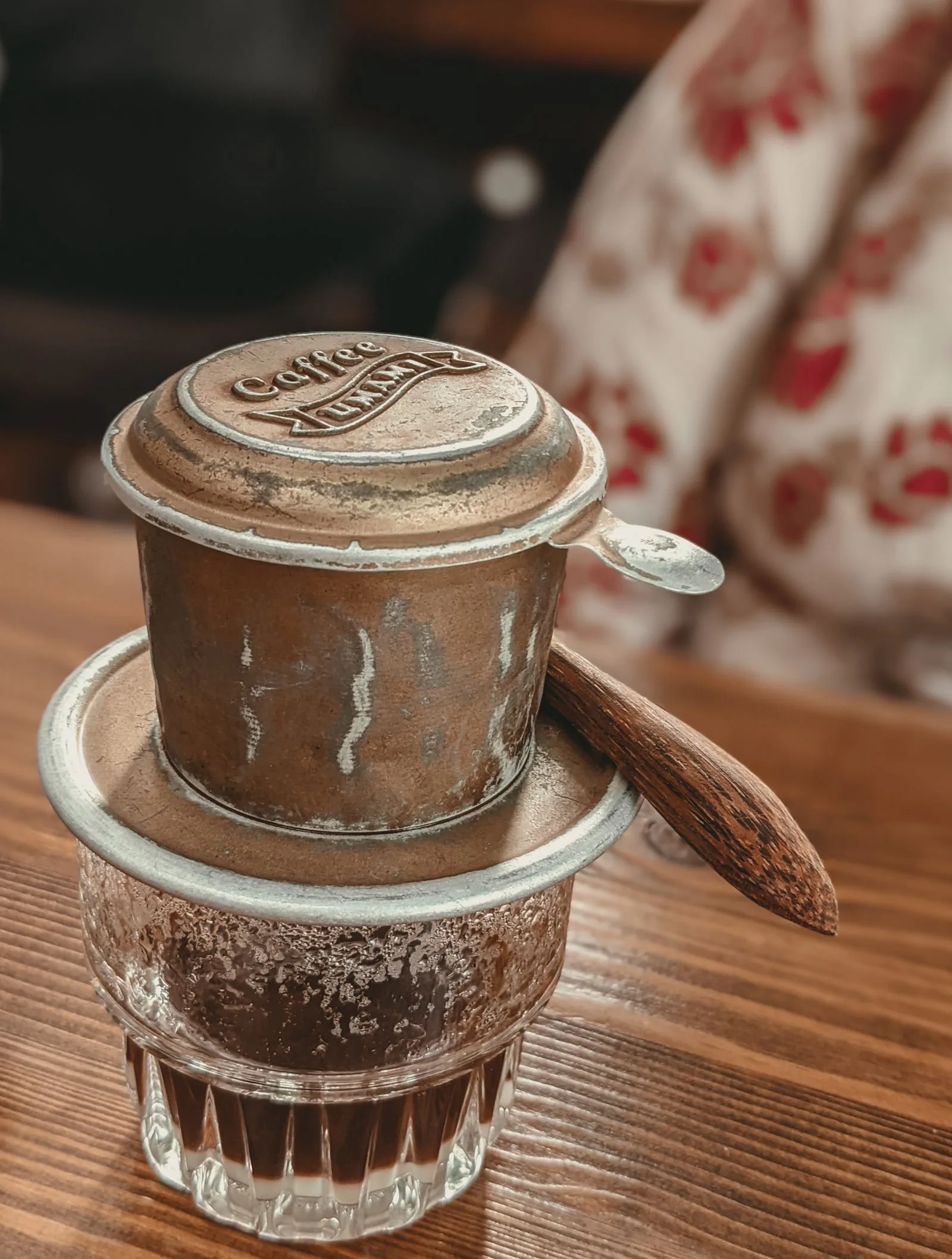 Filtre de métal patiné et taché, couvercle gravé en place, posé sur un verre à facettes où le café a déjà coulé.