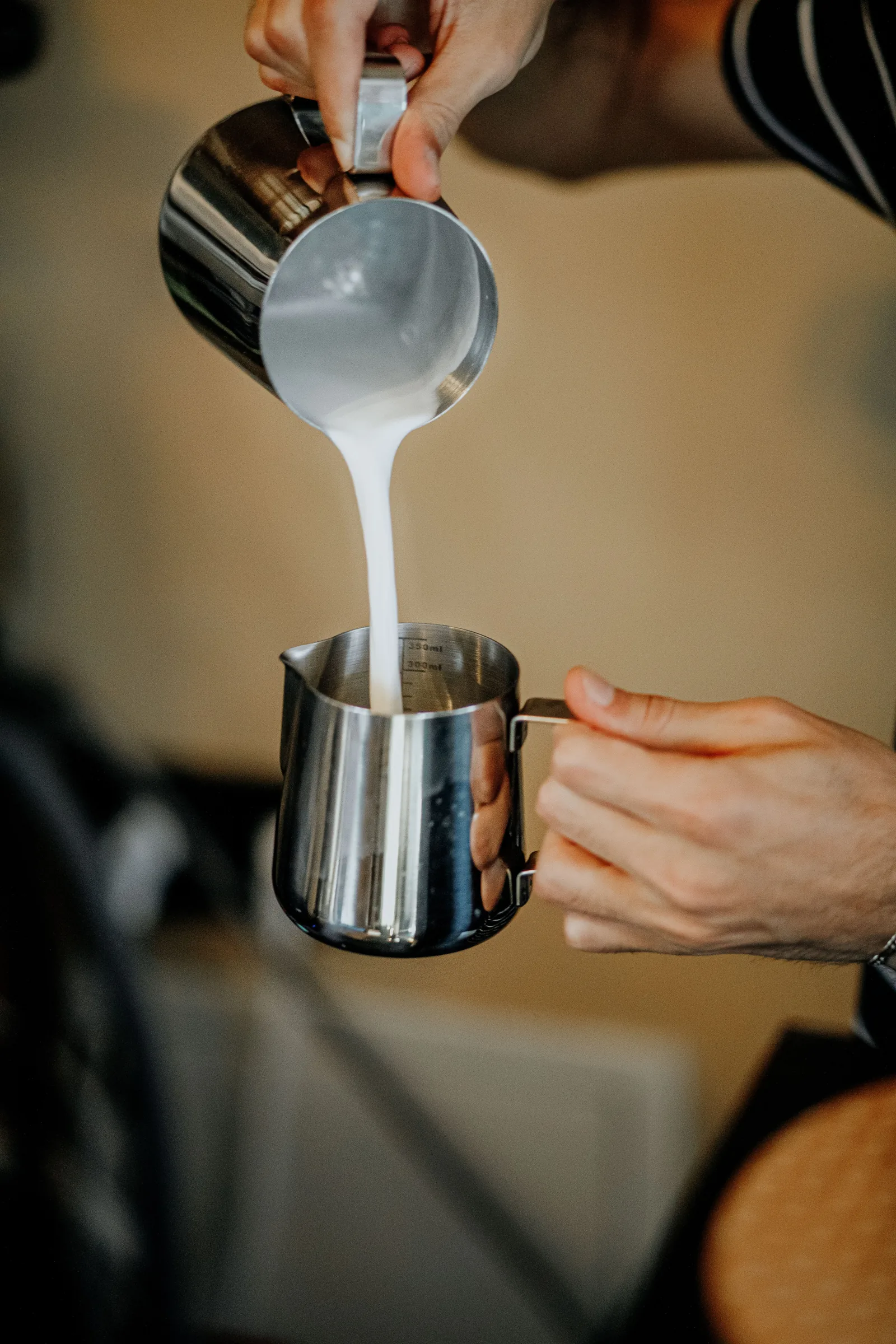 Des mains versent du lait mousseux d'un pichet métallique dans un autre, sans qu'aucun visage ne soit visible