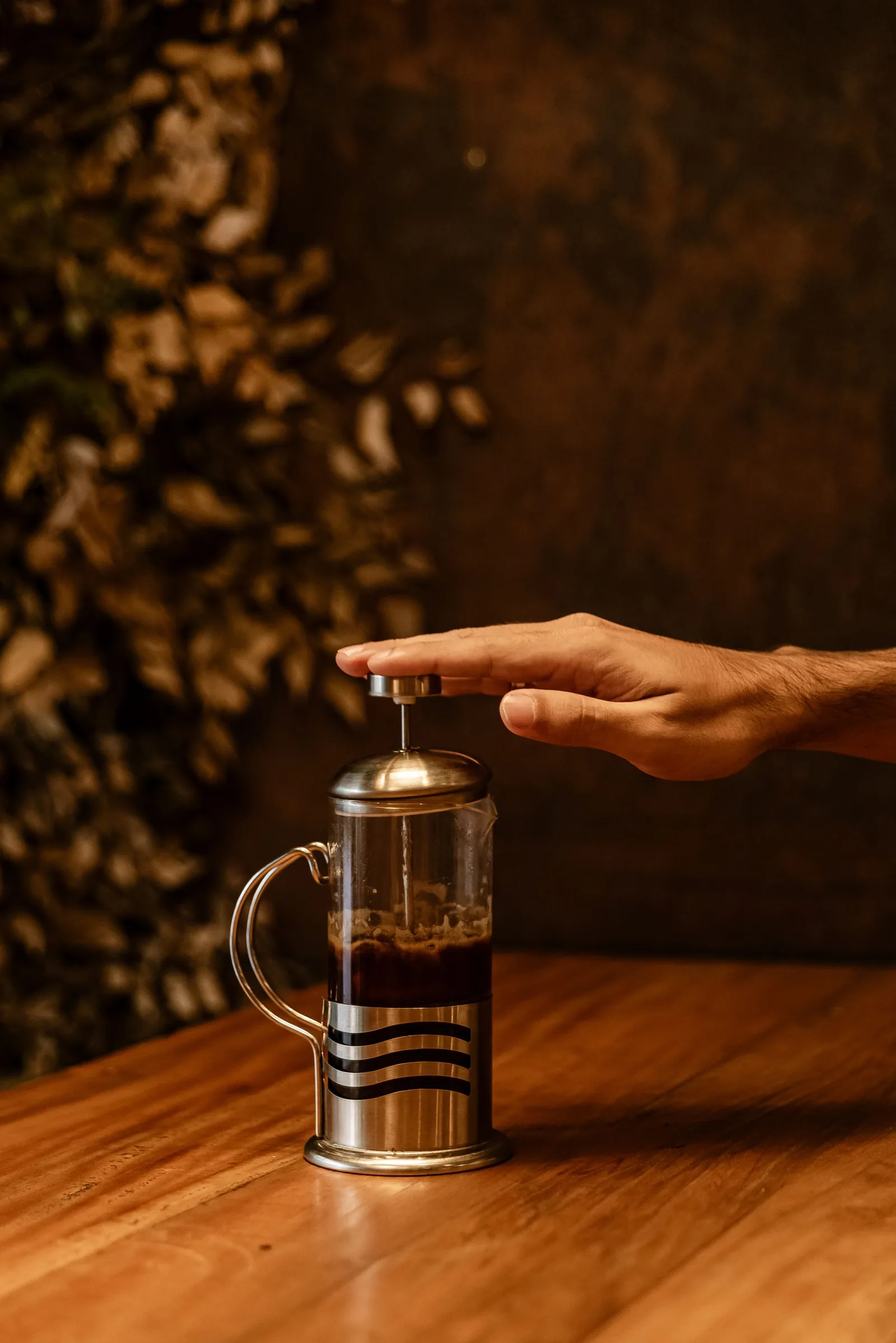 Une main appuie sur le bouton du piston d'une cafetière à piston en verre remplie de café, posée sur une table de bois sombre.