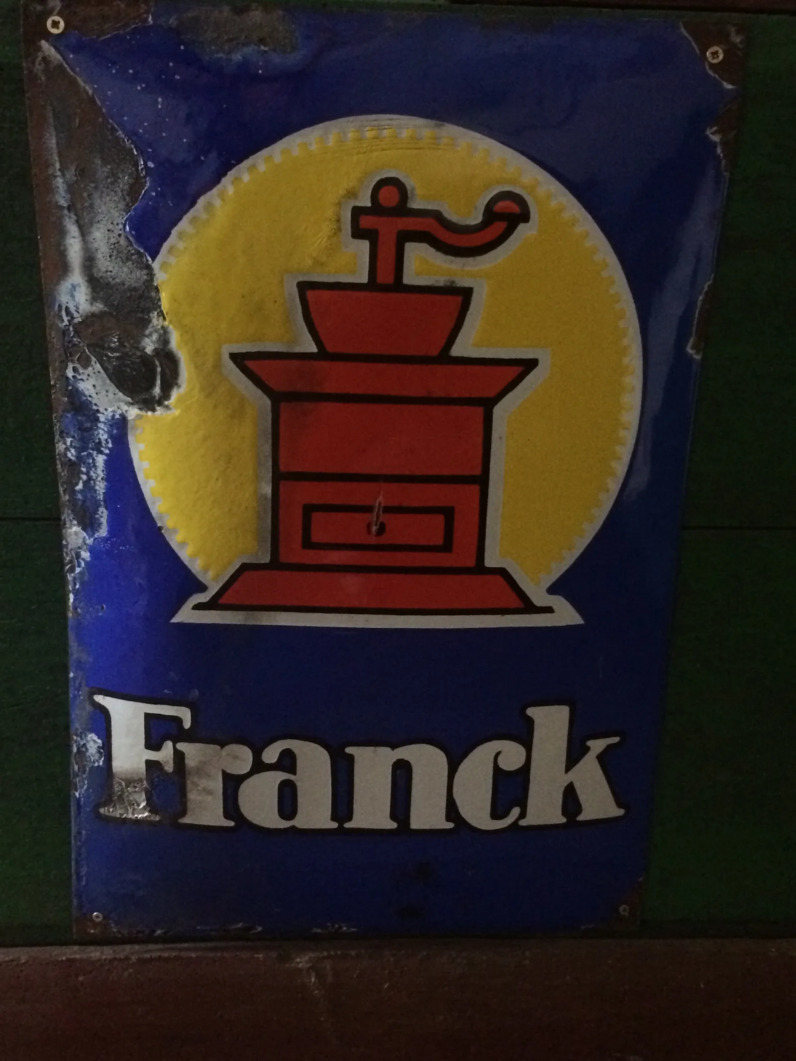 Photographie d'une plaque publicitaire émaillée bleu foncé, écaillée sur les bords, portant un large disque jaune où se détache un moulin à café rouge, avec le nom Franck en grandes lettres claires au bas.