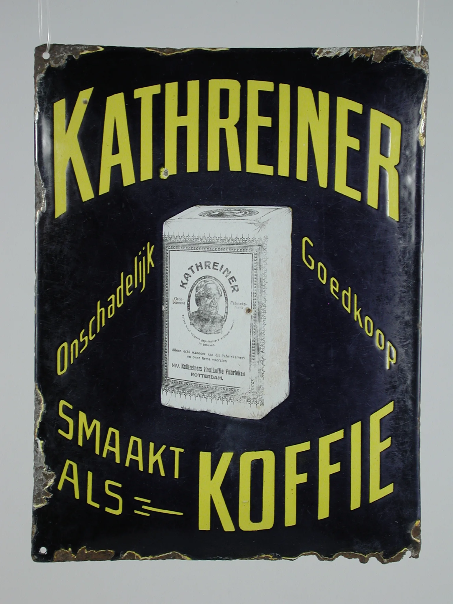 Photographie d'une plaque publicitaire émaillée bleu foncé à lettres jaunes, montrant un paquet de café de malt Kathreiner présenté comme substitut du café.