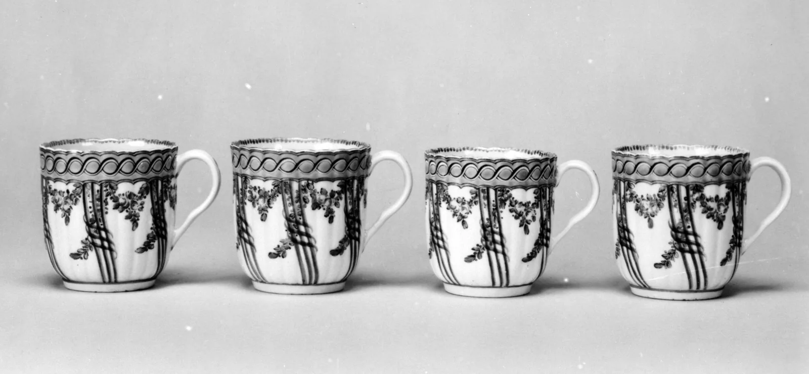 Photographie en noir et blanc de quatre gobelets à café de porcelaine anglaise alignés de face, anses tournées dans le même sens, chacun orné d'une figure peinte en camaïeu sous une frise en haut de paroi.