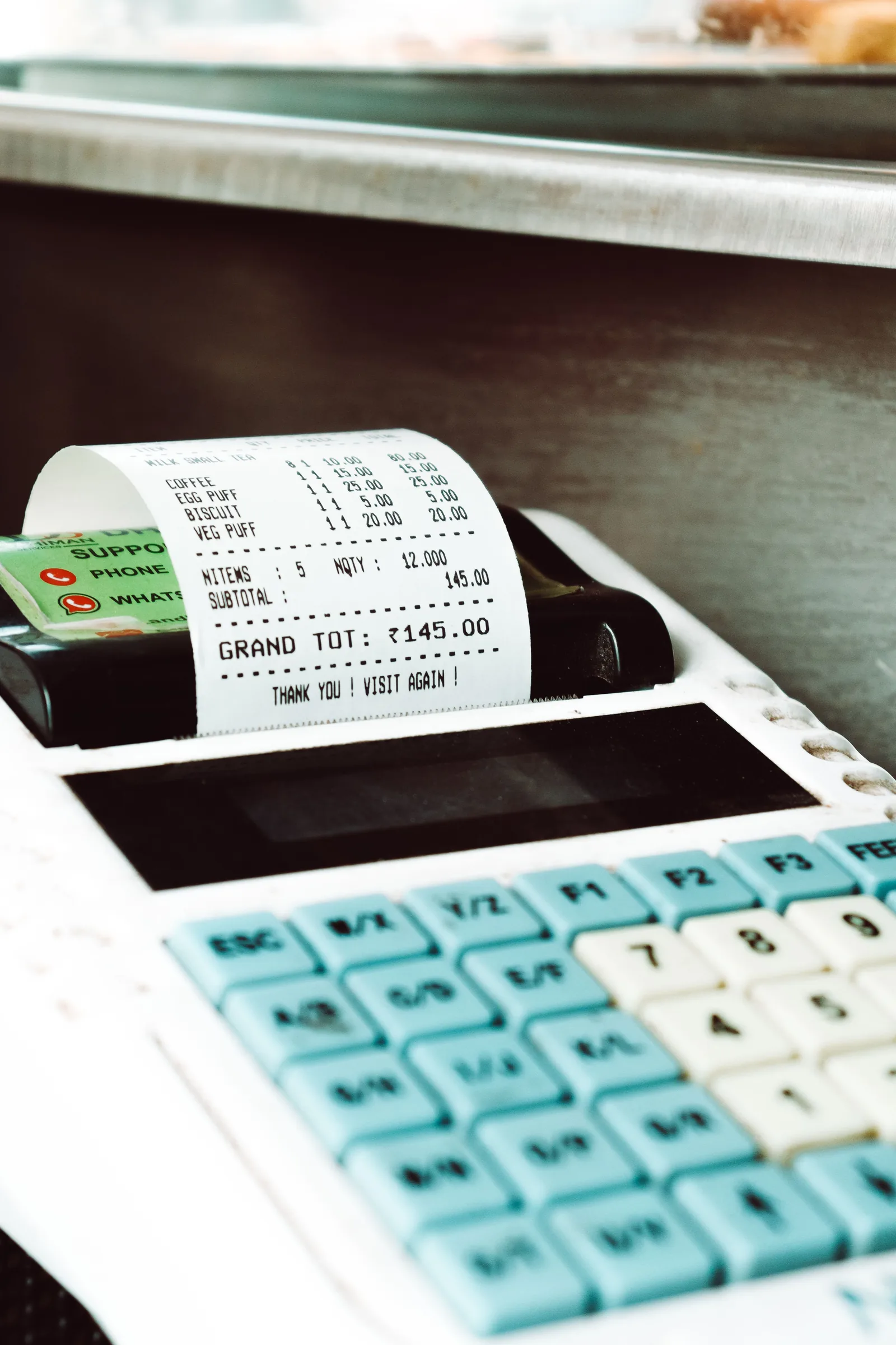 Un ticket de caisse sort d'une caisse enregistreuse à touches turquoise, avec une ligne indiquant un café à 15 roupies parmi d'autres articles vendus