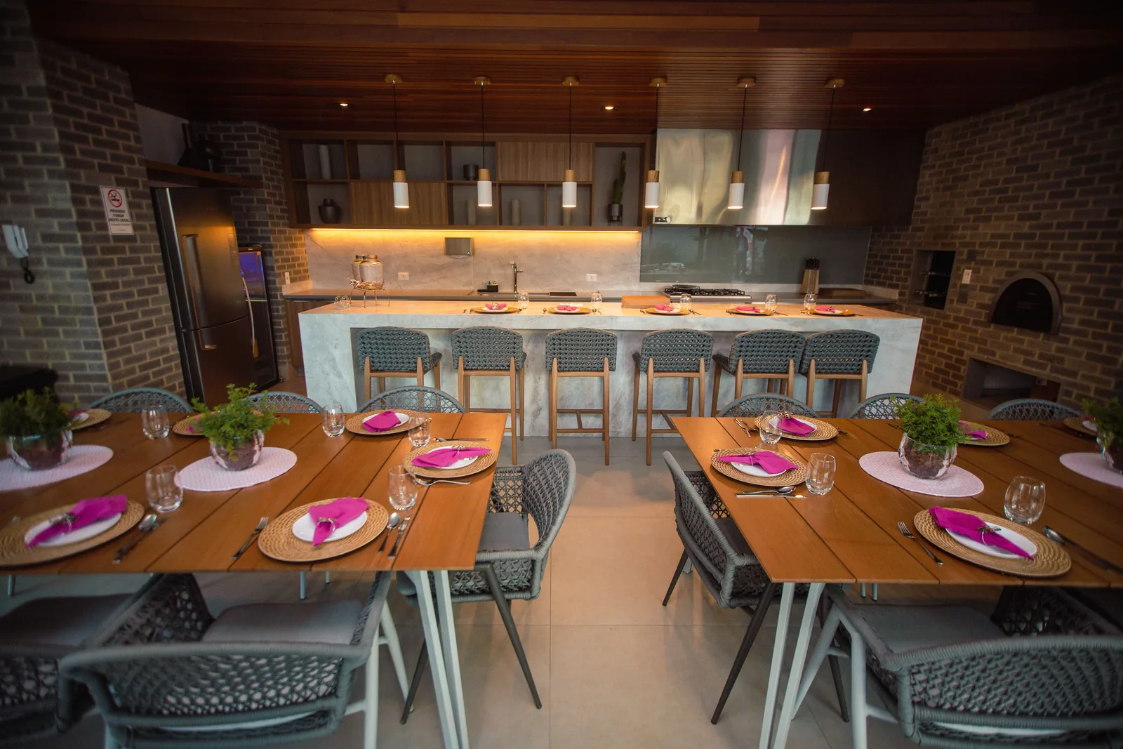 Vue depuis une cuisine ouverte sur une salle de restaurant avec plusieurs tables dressées de couverts et de serviettes roses, un bar et un four à bois en arrière-plan