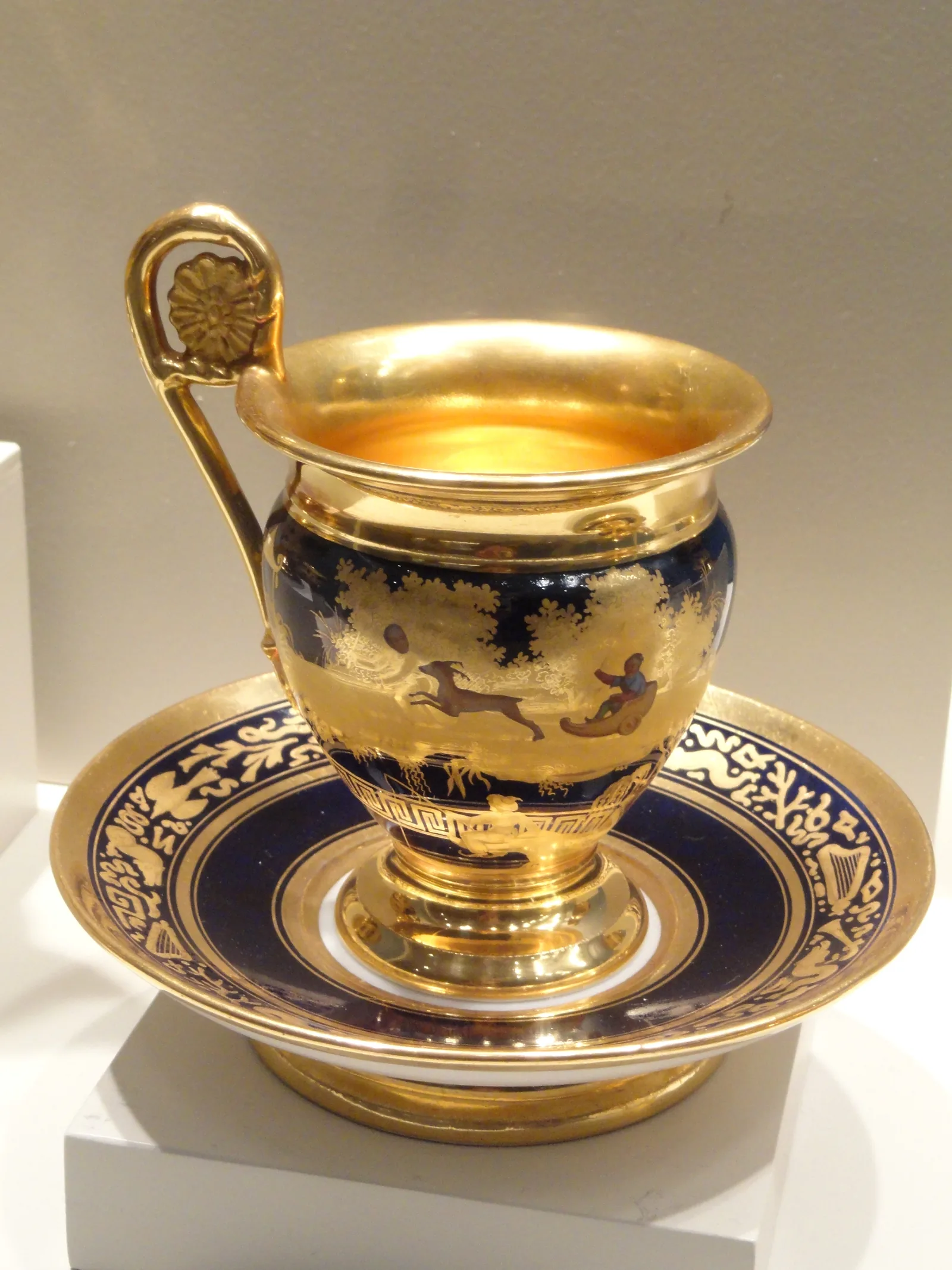Photographie d'une tasse à café de porcelaine parisienne et de sa soucoupe : le corps de la tasse porte une frise dorée sur fond bleu nuit, l'intérieur et l'anse sont entièrement dorés, la soucoupe reprend le même bandeau bleu et or.