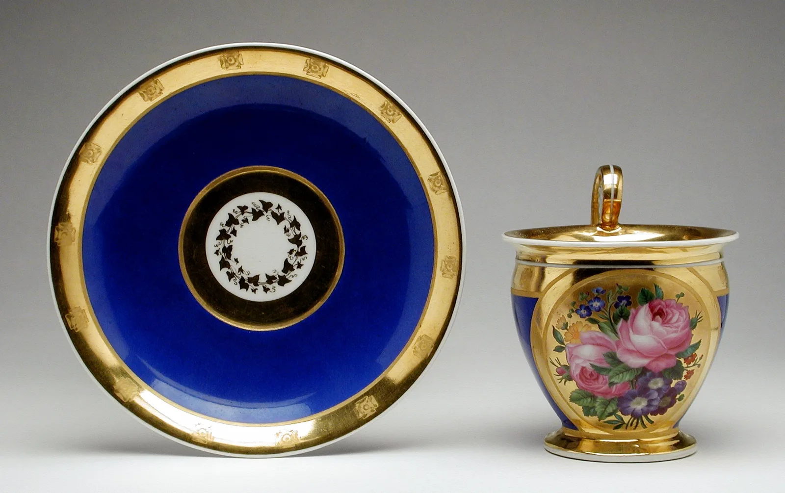 Photographie d'une tasse à café de porcelaine viennoise à fond entièrement doré, décorée d'un bouquet de roses peintes, présentée avec sa soucoupe à large bandeau bleu cobalt cerné d'or.