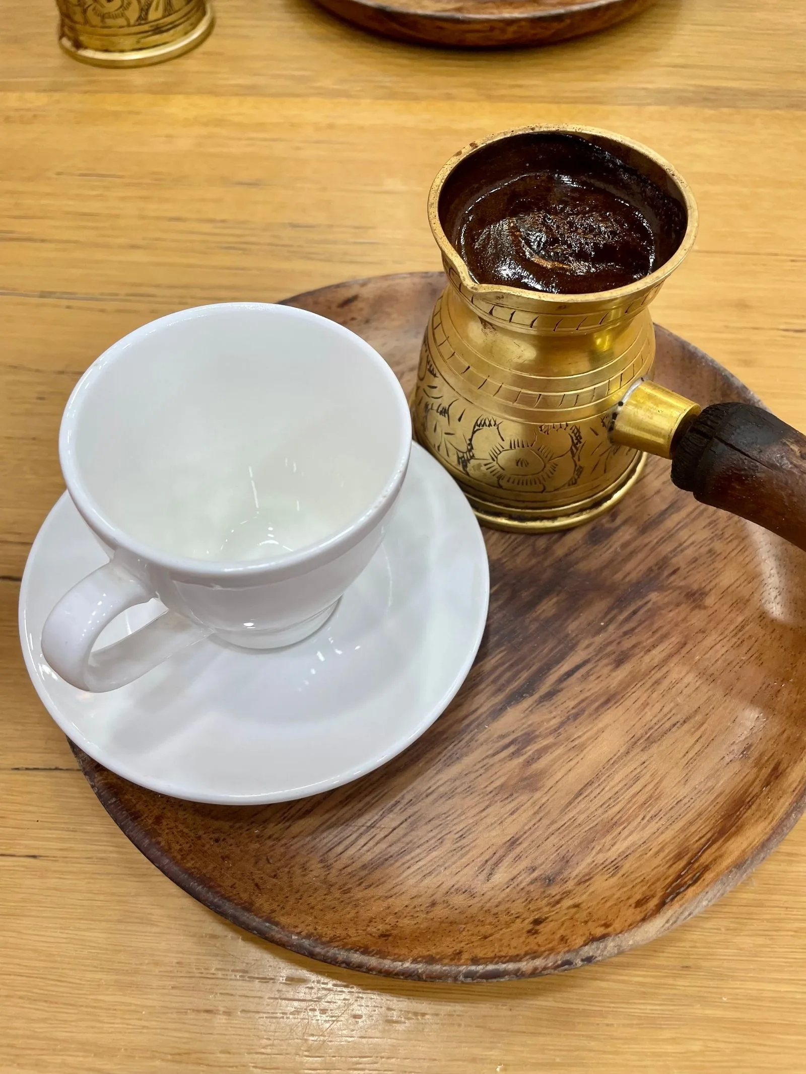 Pot de laiton gravé rempli de café à ras bord et tasse blanche renversée sur sa soucoupe, sur un plateau de bois tourné.
