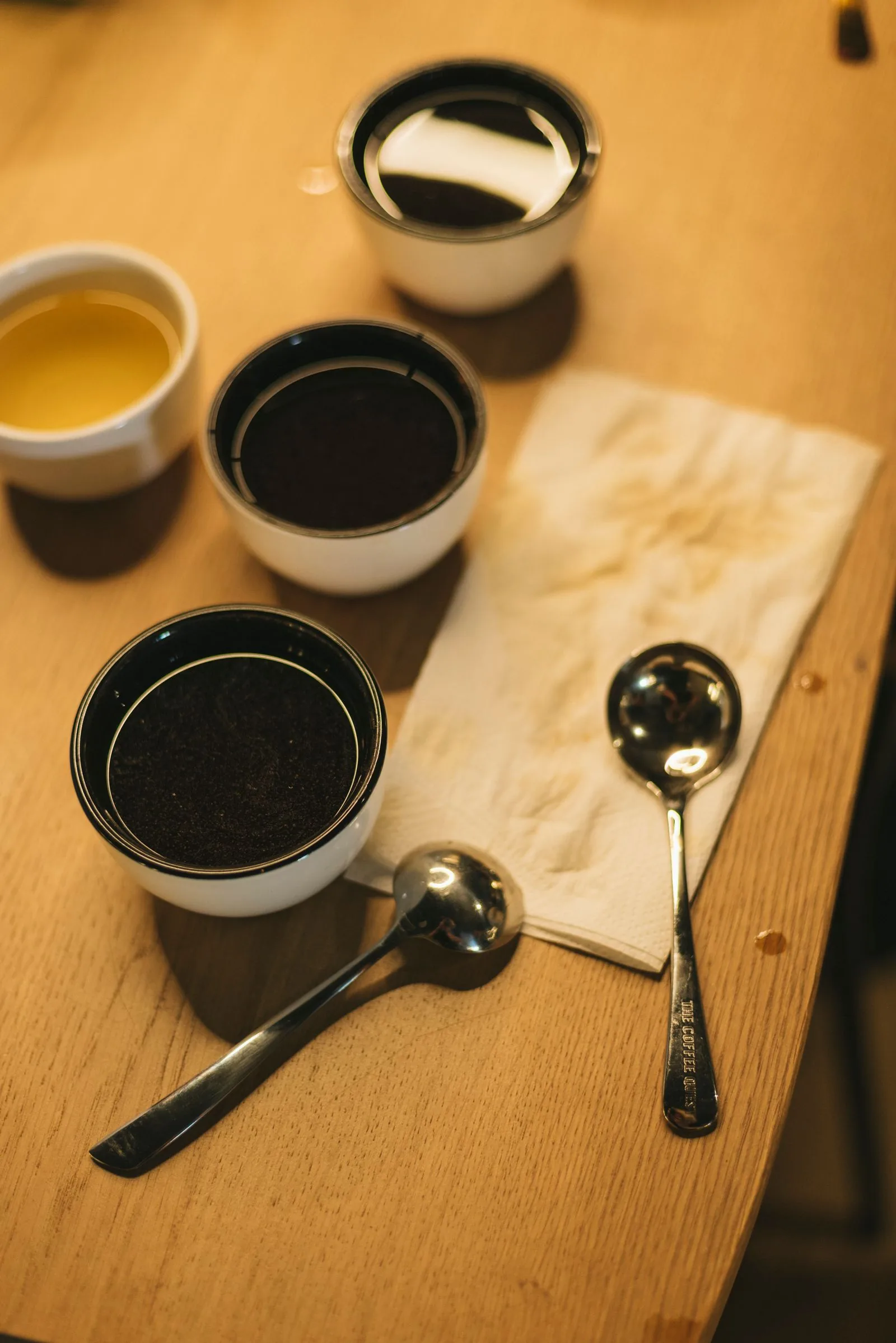 Quatre bols de porcelaine remplis d'infusions de teintes différentes, deux cuillères de dégustation et une serviette de papier sur une table claire.