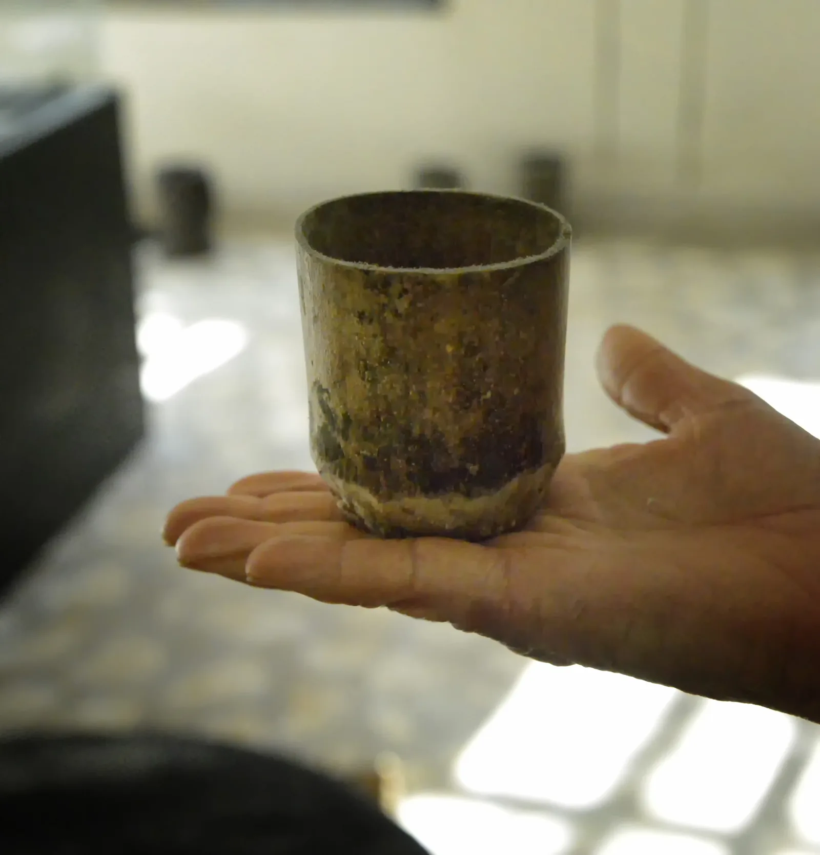 Photographie rapprochée d'un petit gobelet à café ancien, sombre et patiné, posé au creux d'une main ouverte, devant une fenêtre en contre-jour.
