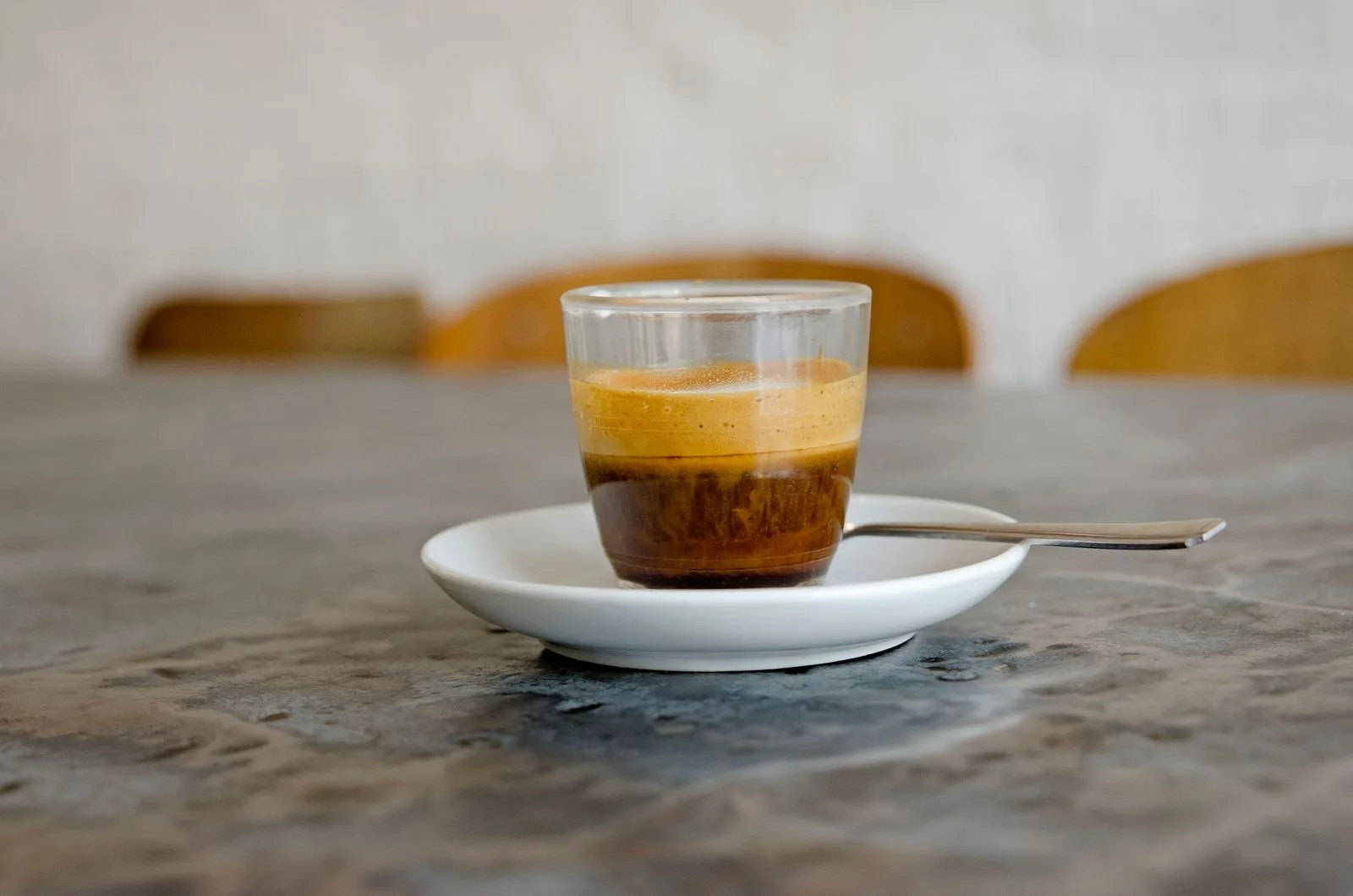 Verre droit contenant un espresso à crème claire, posé sur une soucoupe blanche avec une cuillère, sur une table de pierre.