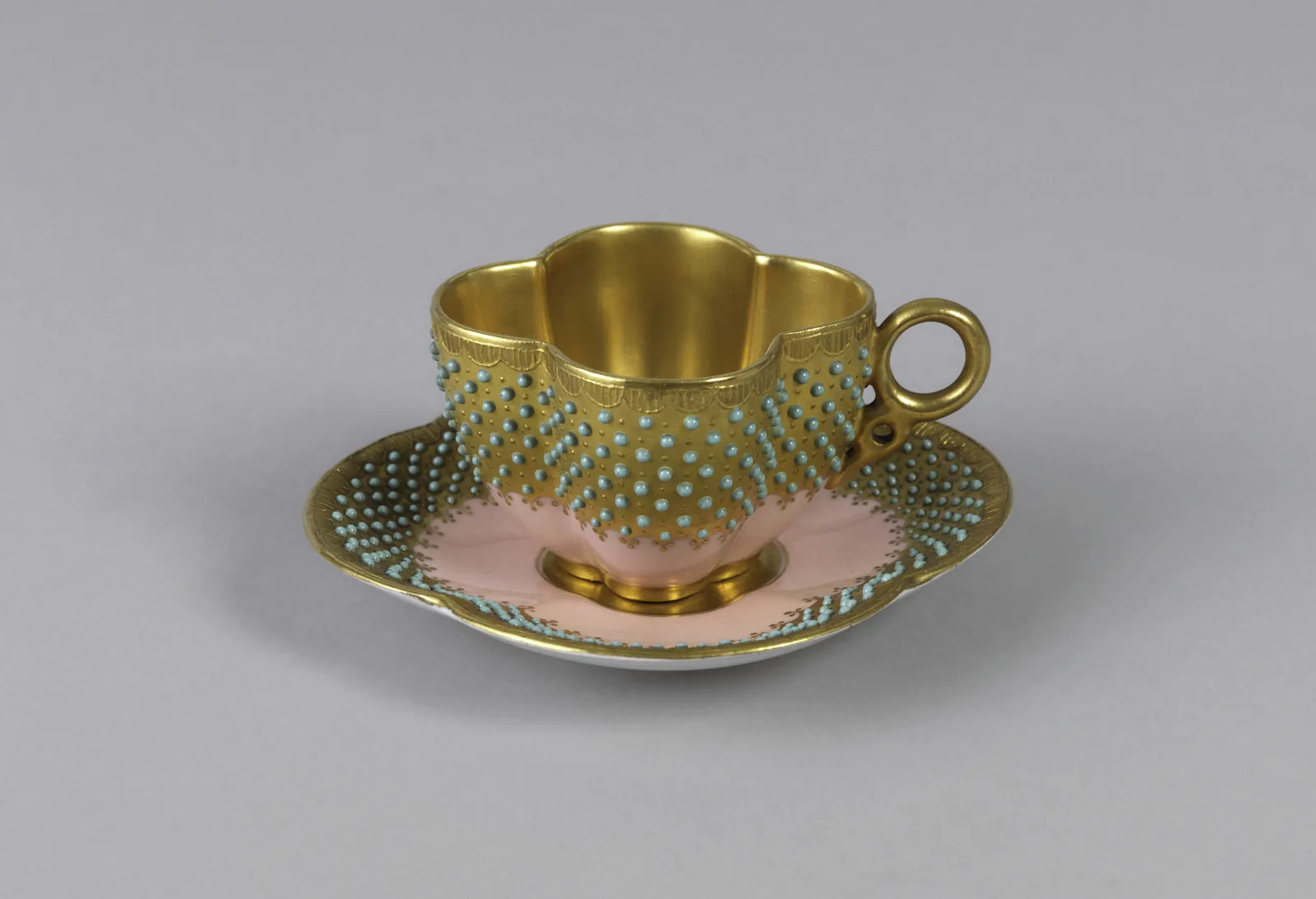 Photographie d'une petite tasse à café quadrilobée et de sa soucoupe assortie, fond rose ceinturé d'un large bandeau d'or ponctué de points verts en relief.