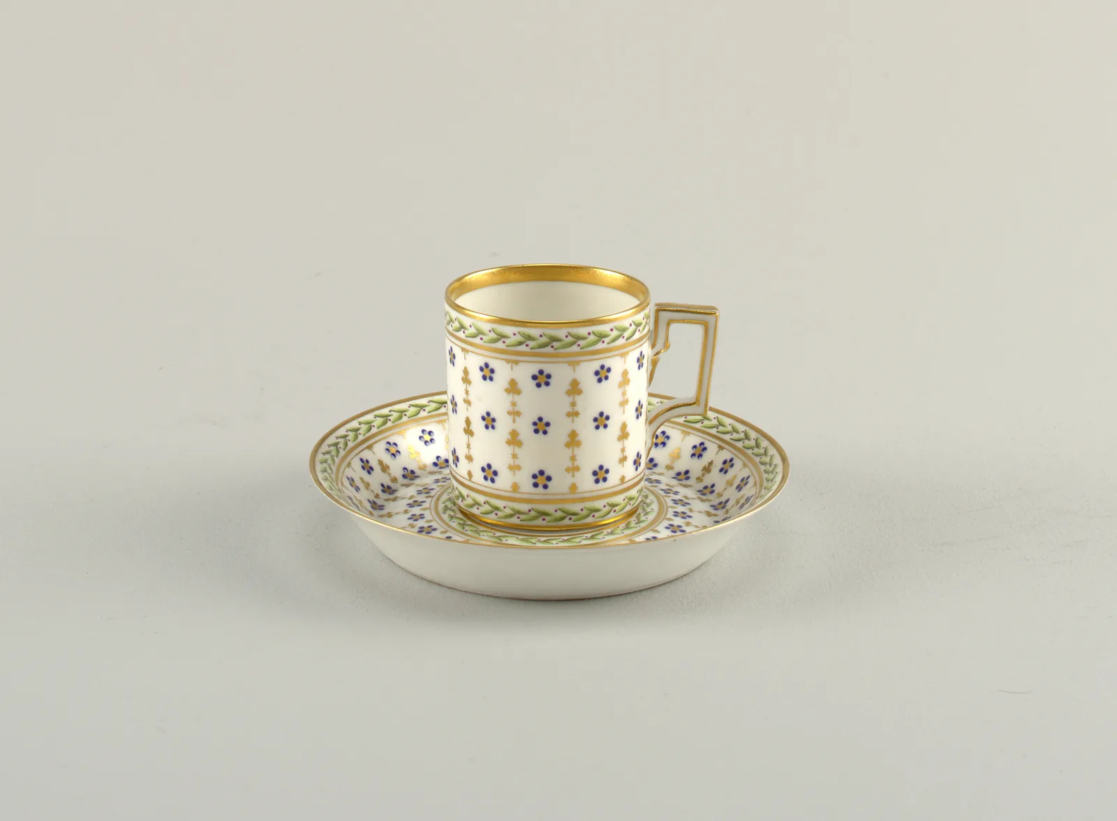 Photographie d'une petite tasse à café de porcelaine et de sa soucoupe, semées de motifs bleus en étoile sur fond blanc et bordées de larges filets d'or, l'anse étant de forme carrée.