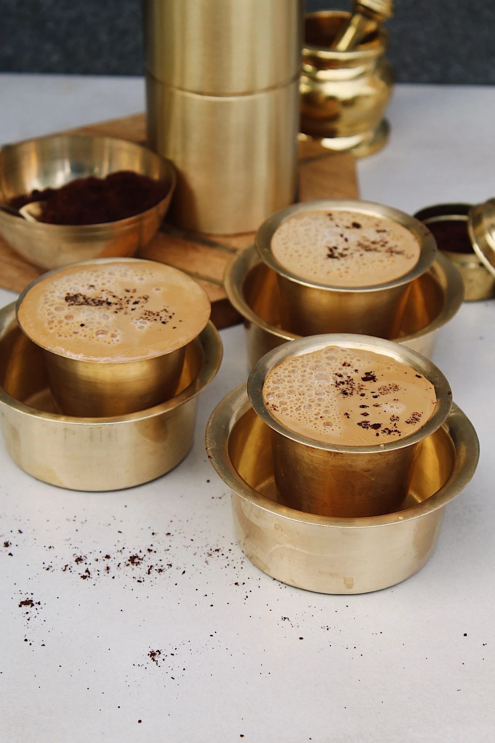 Trois gobelets de laiton posés chacun dans leur coupelle, remplis d'un café mousseux, grains moulus renversés sur la table claire.