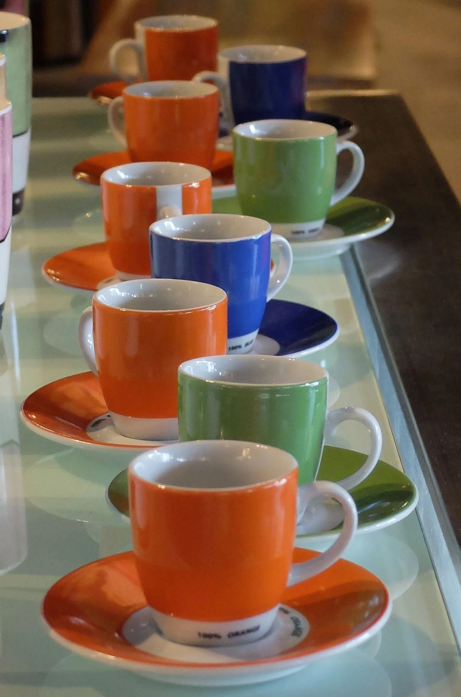 Rangée de tasses orange, bleues et vertes posées sur leurs soucoupes assorties, le long d'une vitrine de comptoir.