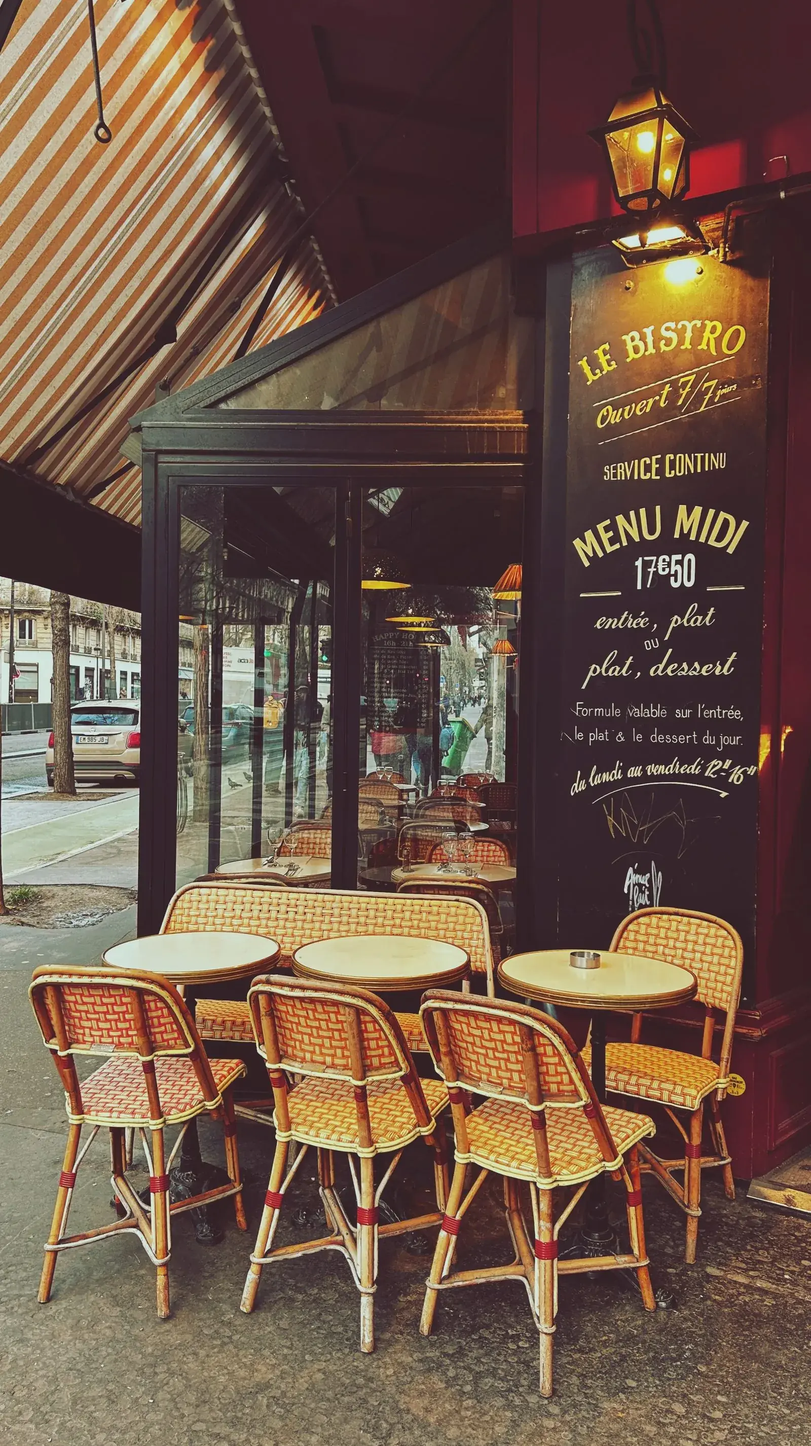 Tables rondes et chaises cannées sur un trottoir, vitrine noire portant « LE BISTRO » et le menu de midi écrit à la craie, lanterne allumée.