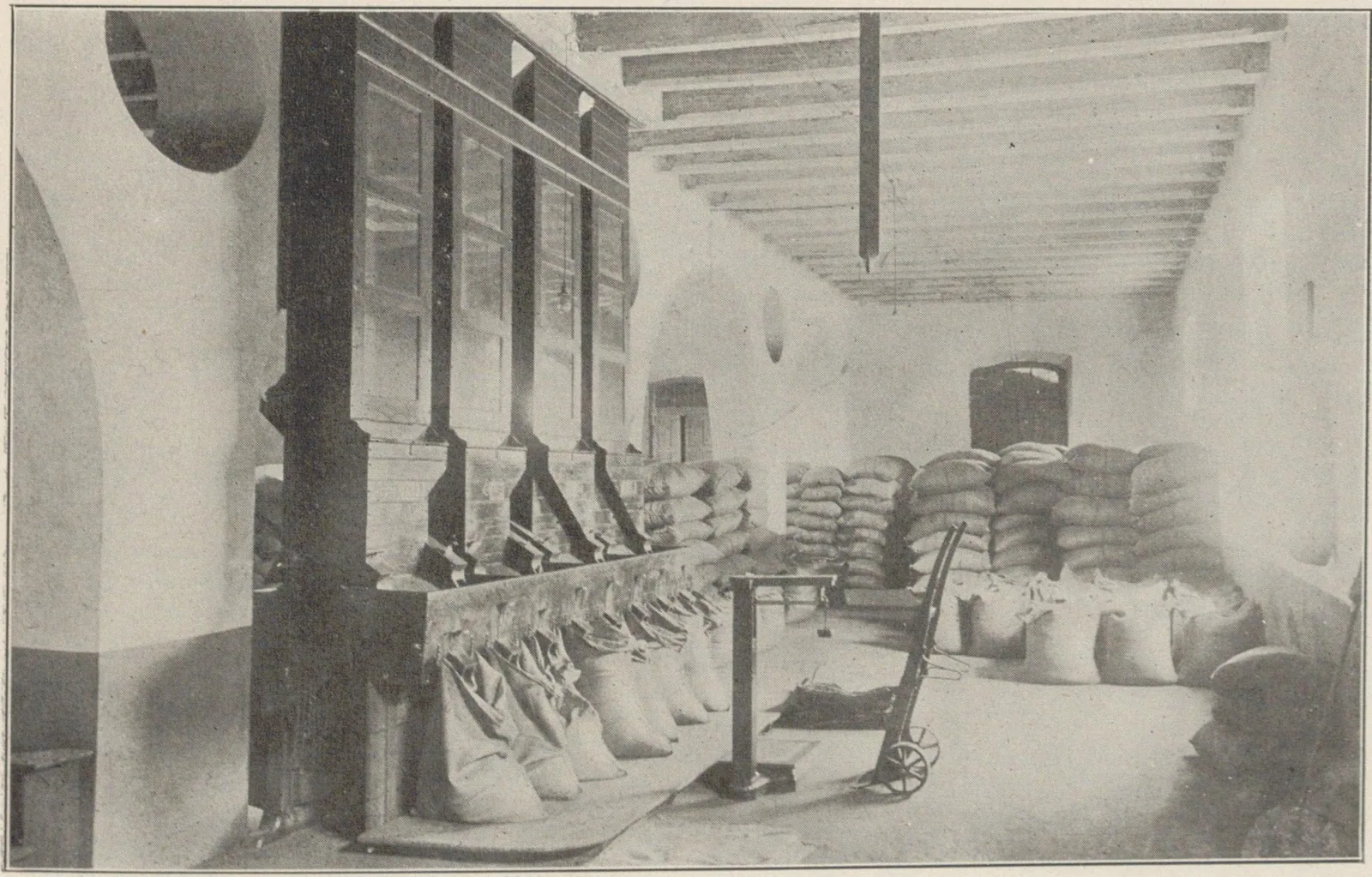 Halle où des sacs pleins sont alignés sous des goulottes de remplissage, à côté de piles de sacs cousus.
