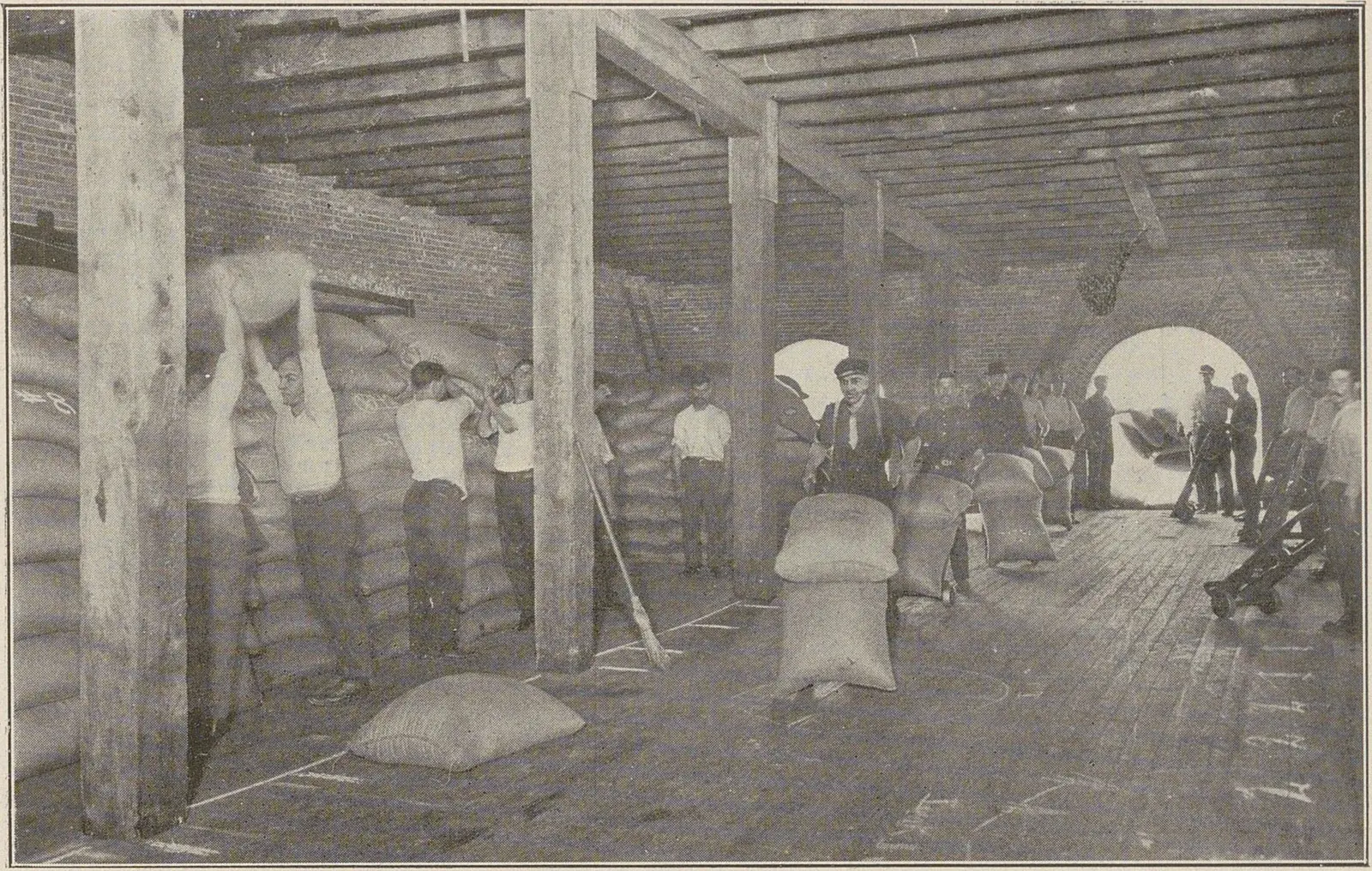 Entrepôt de bois où des hommes portent des sacs de café sur l'épaule entre des piles rangées par marques.