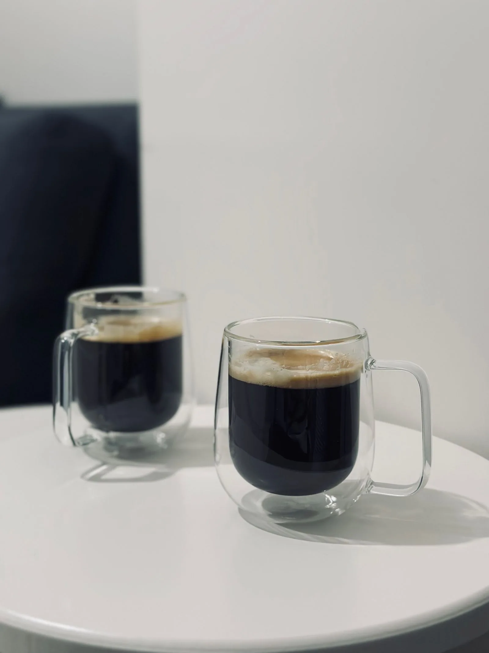 Deux tasses de verre à double paroi avec anse, remplies de café noir, posées sur un guéridon blanc rond.